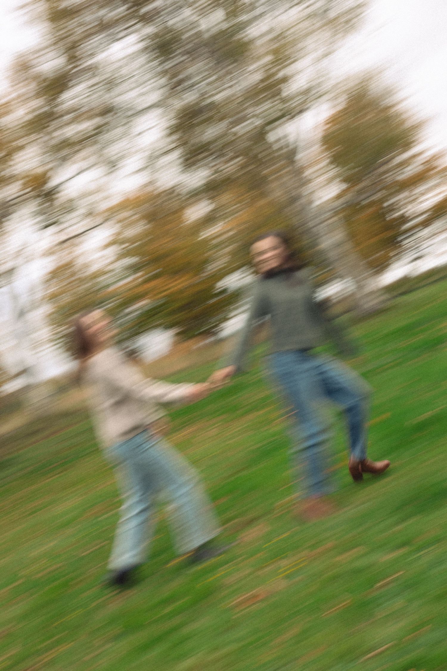 Motion-blurred image of two women holding hands and running across a grassy hillside beneath autumn trees on an overcast day.