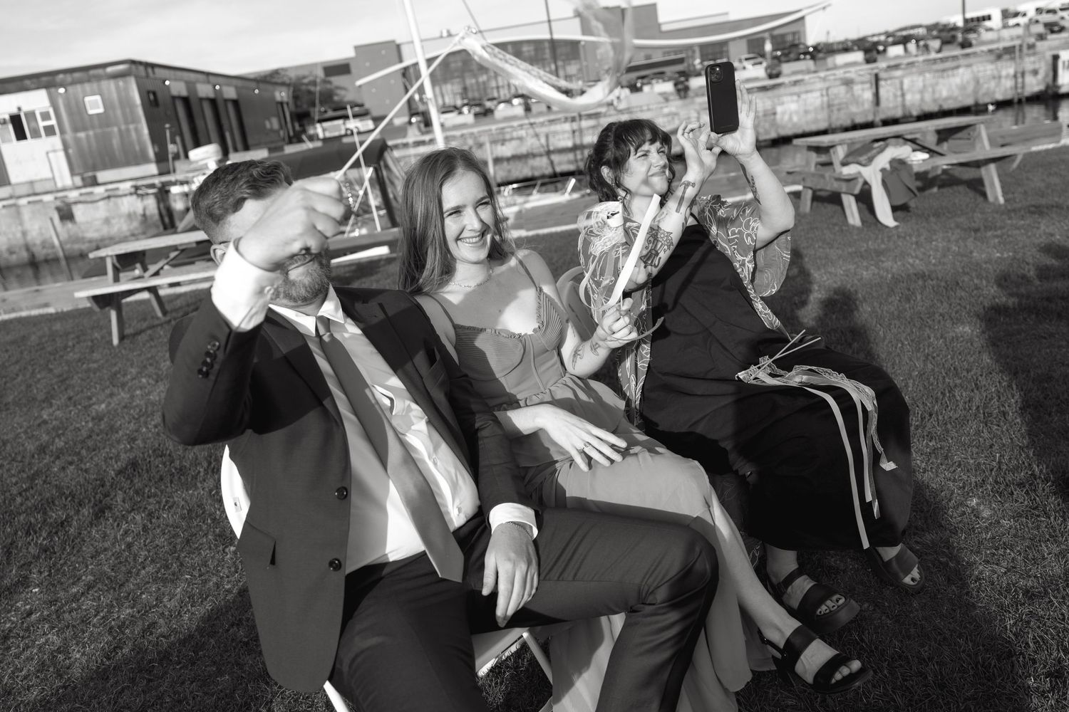Three people in formal attire sit on a grassy lawn near a waterfront, laughing and toasting with drinks in a black and white photo.