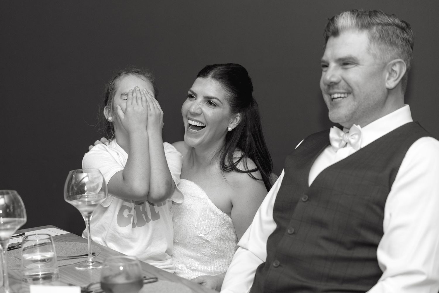 Black and white photo of a laughing bride in a wedding dress beside a smiling groom in a vest and bow tie, with a child covering her face.