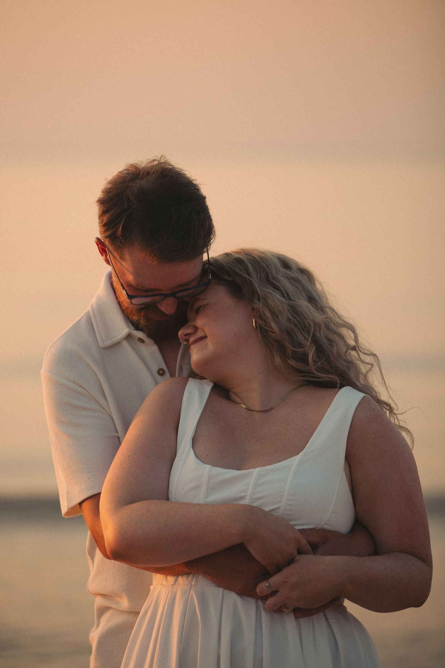 Couple embracing at sunset on a beach, woman in white dress smiling as man in white shirt holds her close.