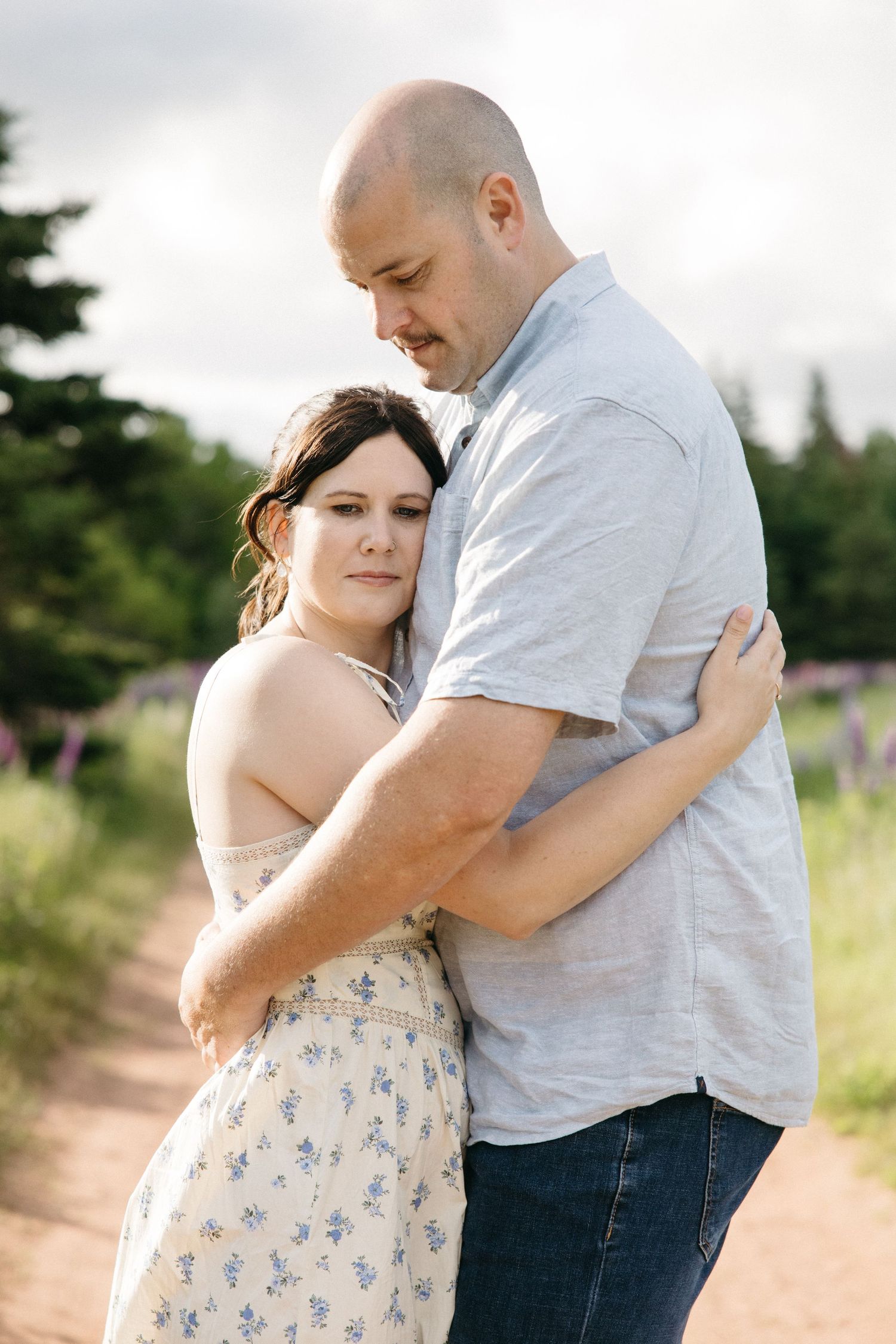 A couple embraces tenderly on a dirt path surrounded by wildflowers and evergreen trees.
