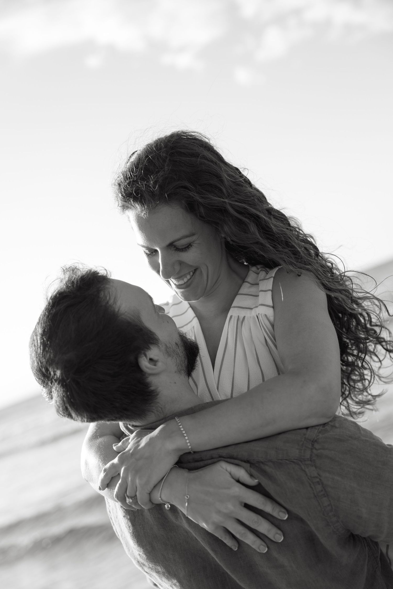 Black and white photo of a couple embracing on a beach, woman laughing as man lifts her up.