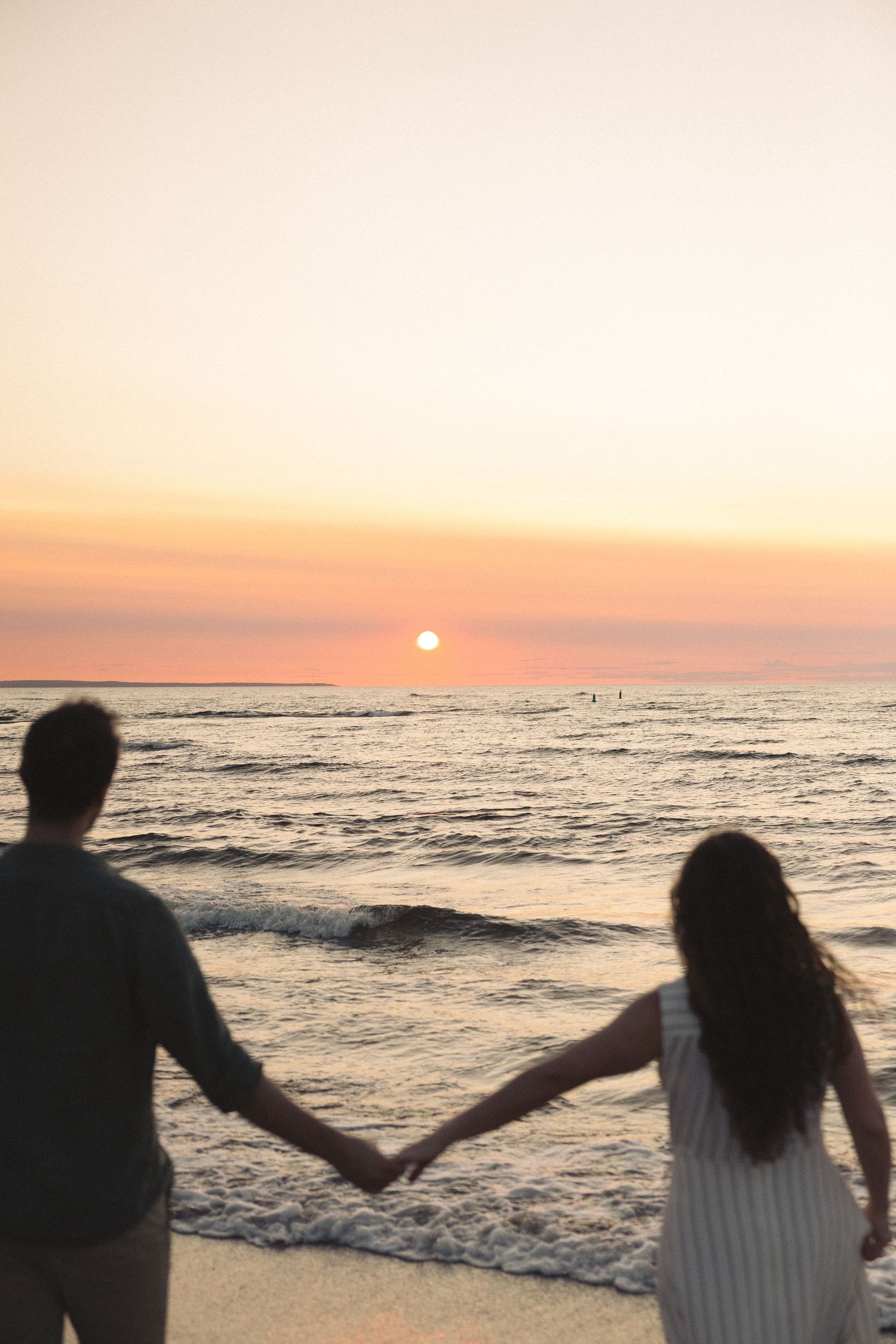 Romantic couple holding hands silhouetted against stunning pink and orange sunset over calm ocean.