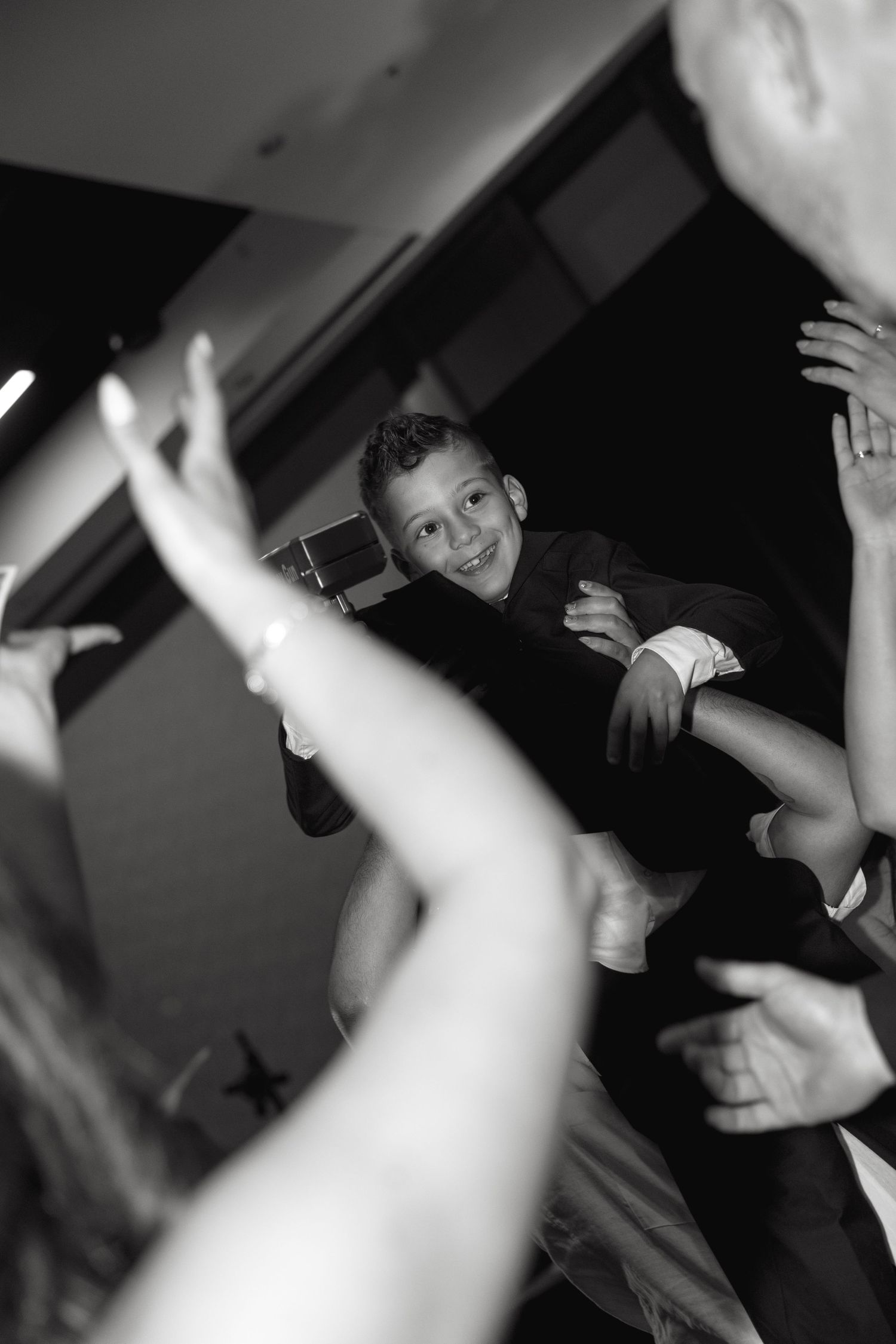 A smiling boy being lifted by adults at a celebration, captured in black and white photography.