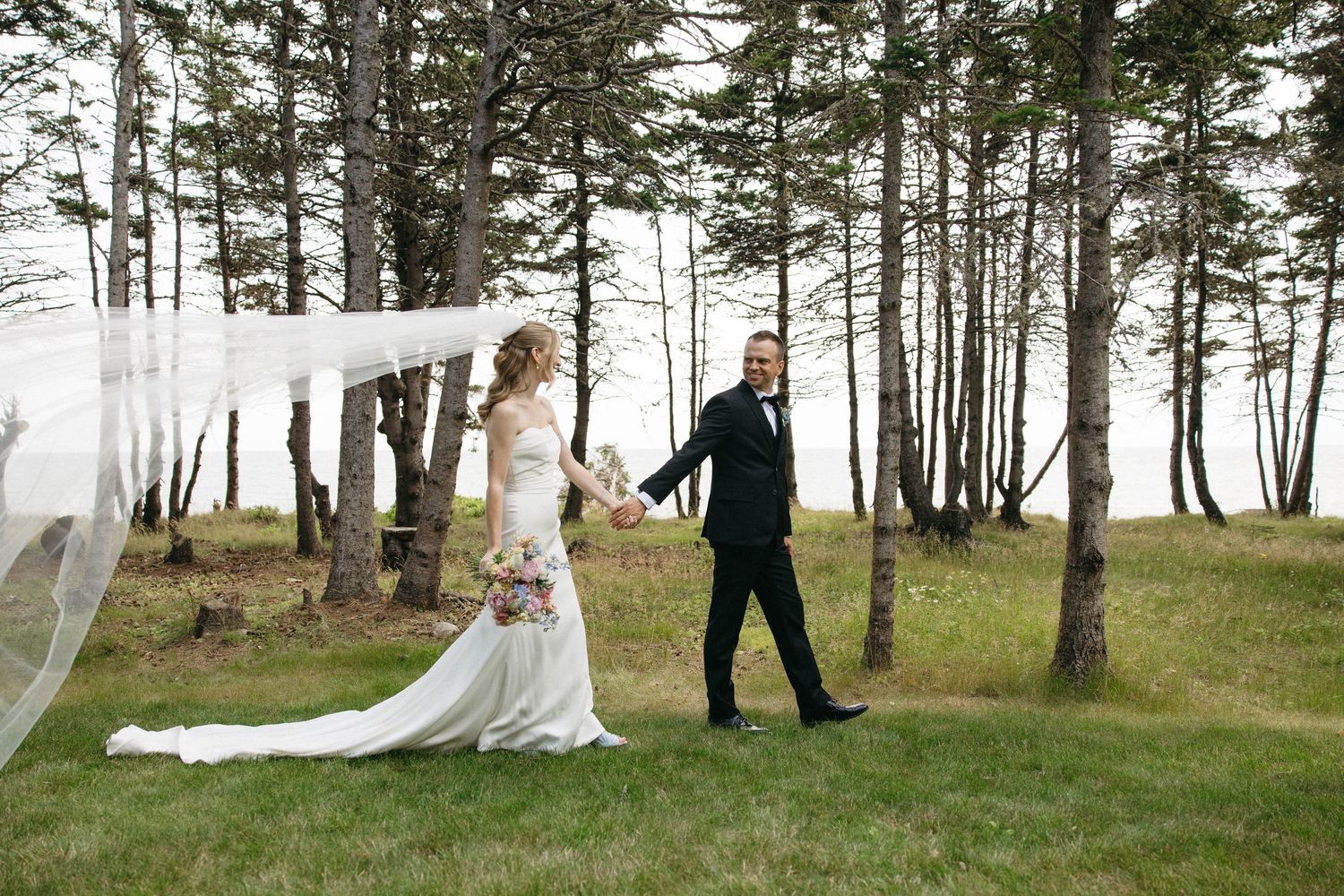 Bride and groom walk hand-in-hand through a lush green forest, her veil flowing dramatically in the wind.
