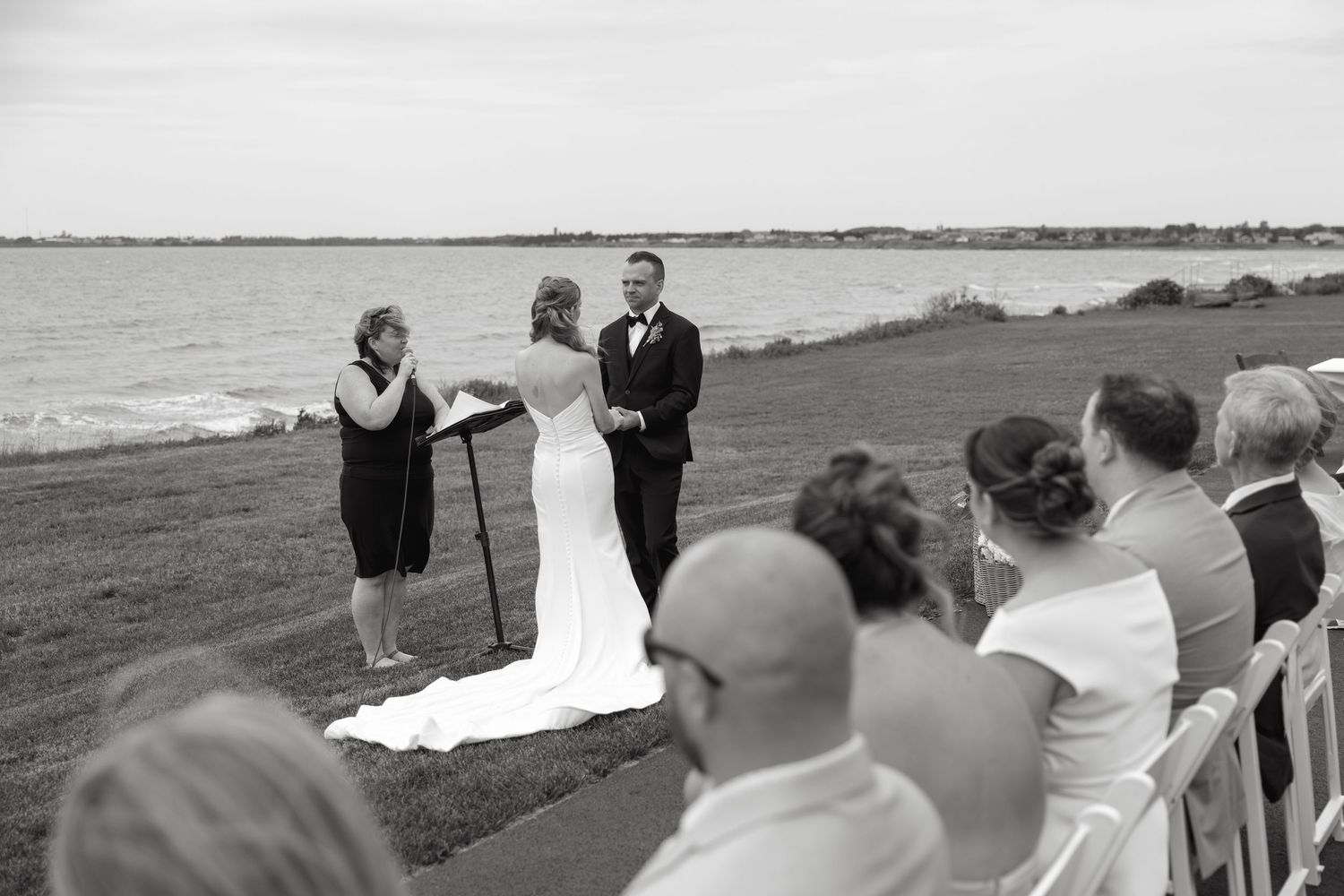 Outdoor waterfront wedding ceremony with bride and groom exchanging vows before seated guests in black and white.