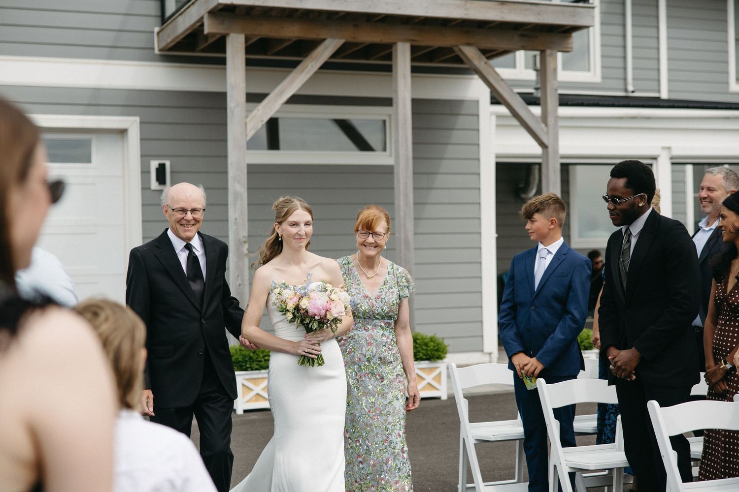 Bride walks down outdoor aisle in white gown holding pink bouquet, escorted by family at a garden wedding ceremony.