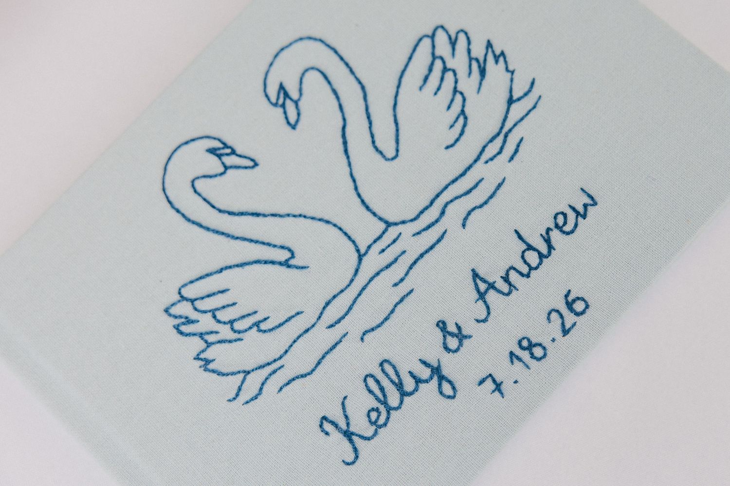 Embroidered swan design on light blue fabric with text reading Kelly and Andrew 7.18.26 in blue thread.