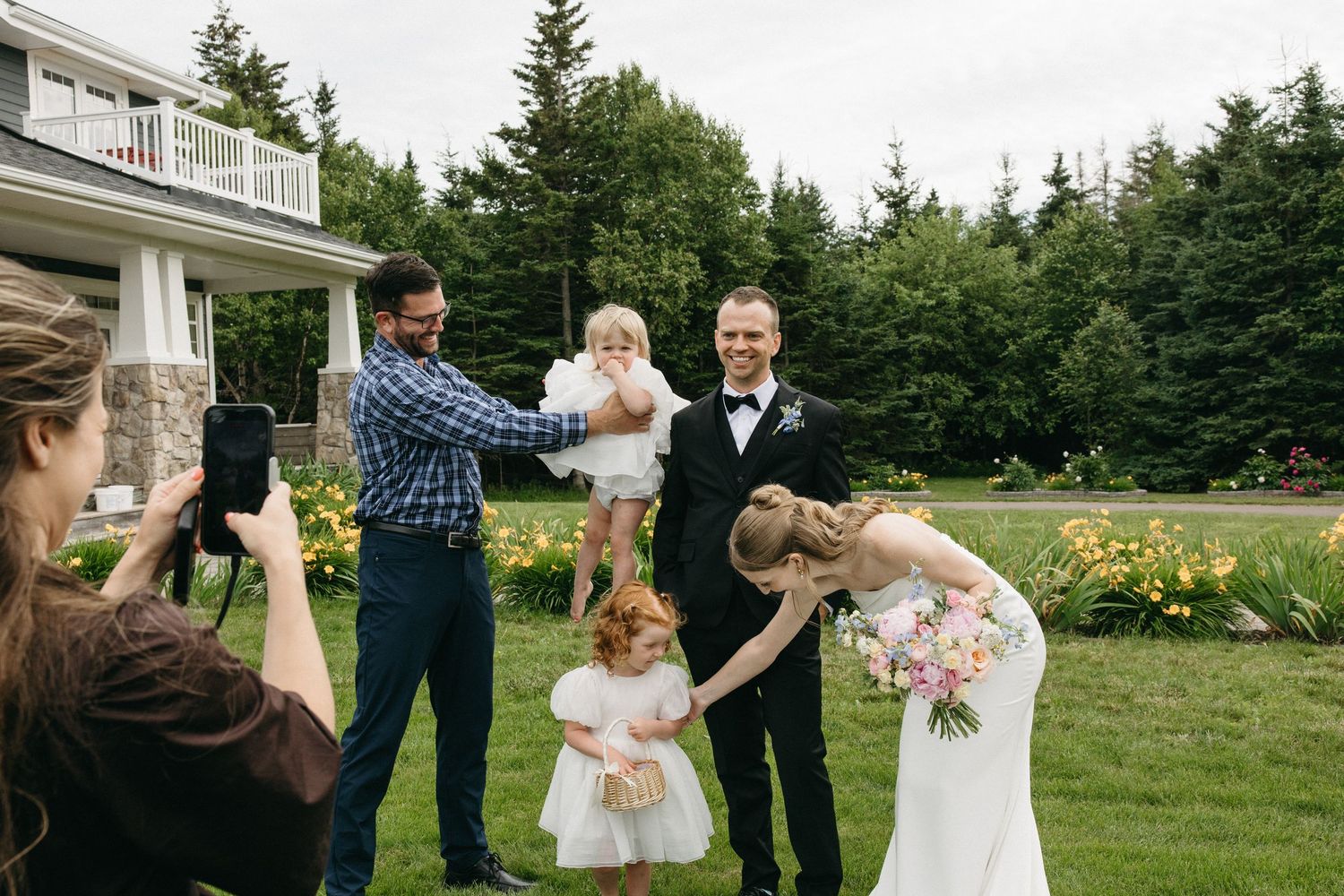 Bride and groom pose with family including young children on green lawn outside elegant home during wedding celebration.