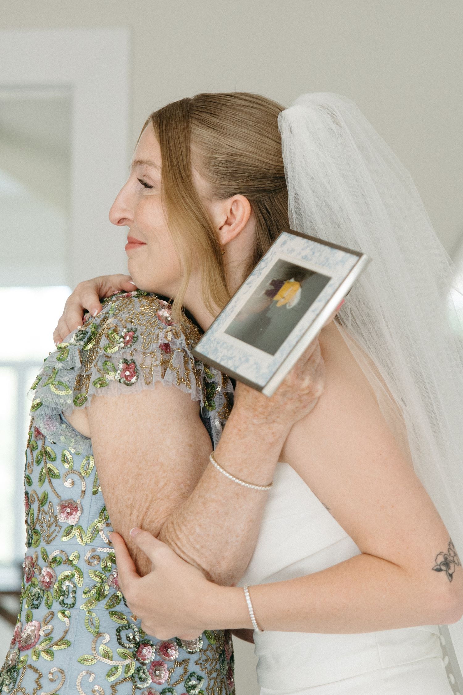 Bride in white gown and veil embracing a woman in floral dress while holding a framed photo.