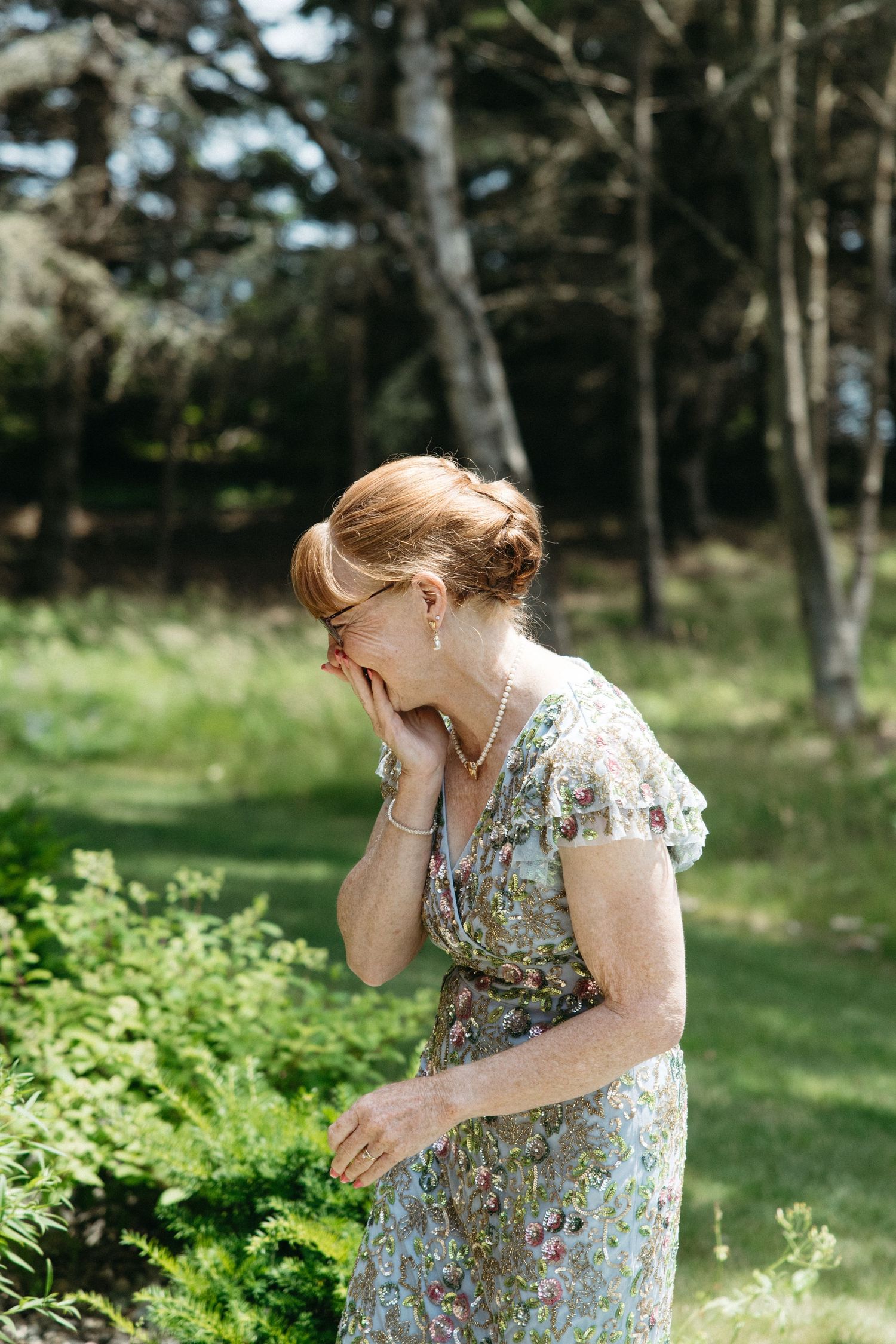 A red-haired woman in a floral dress laughs while covering her face with her hand in a sunny garden.