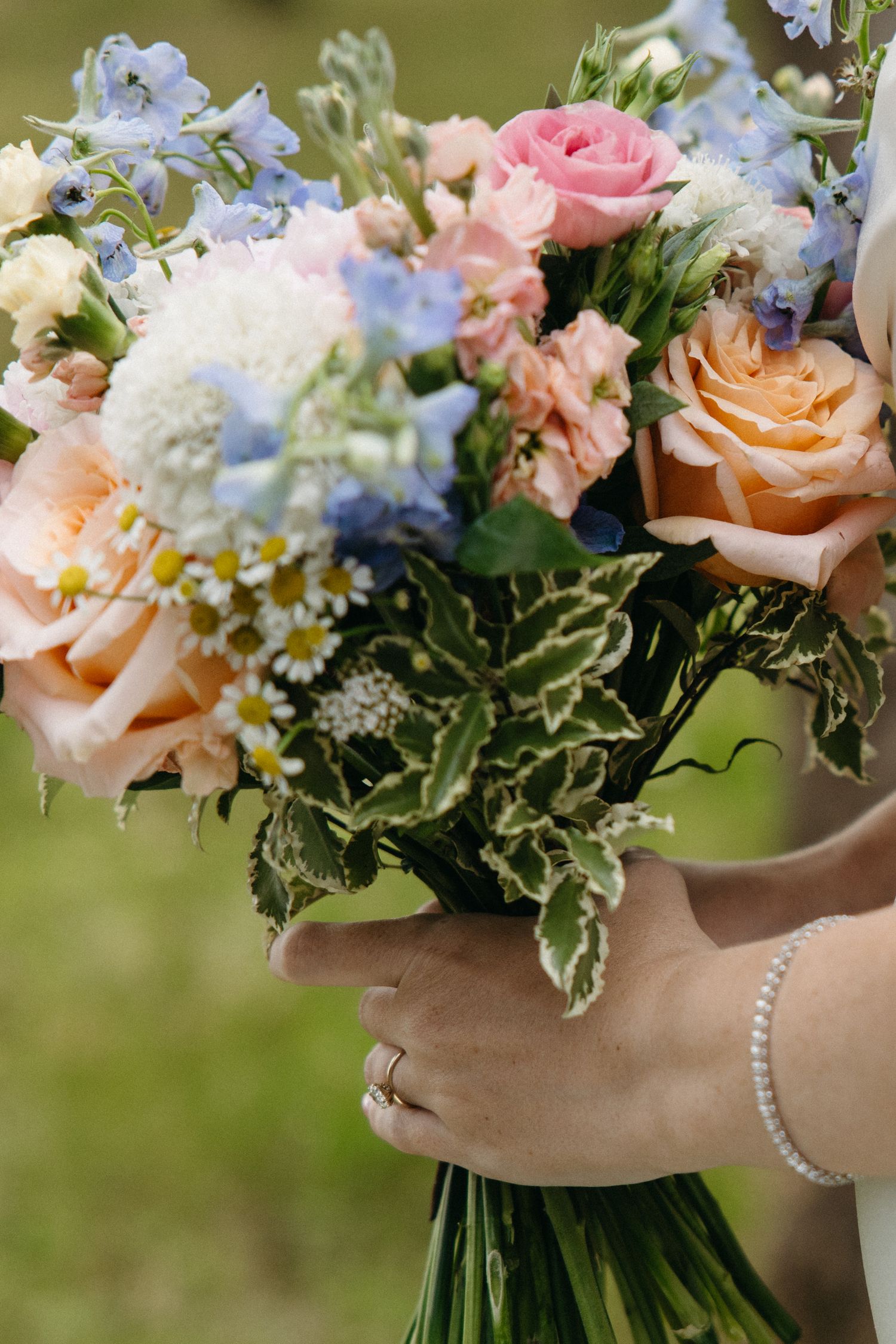 Close-up of bridal bouquet featuring peach roses, pink blooms, blue delphiniums, and daisies.
