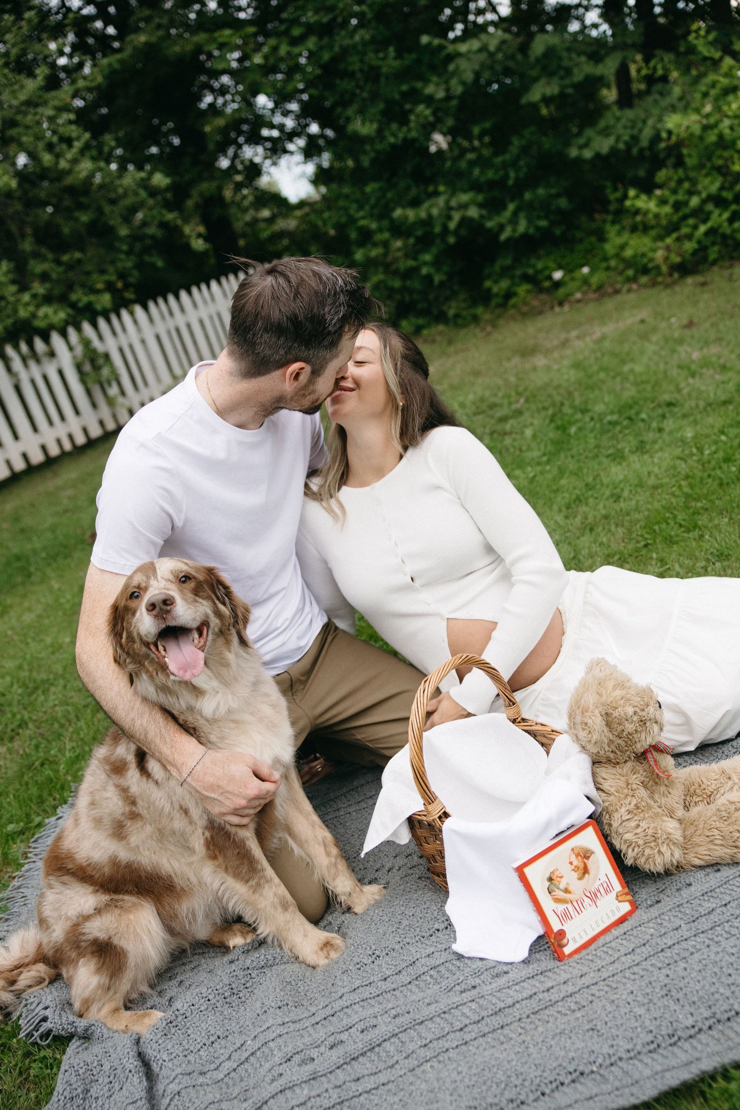 Couple kissing on picnic blanket with their dog, basket, and teddy bear outdoors.