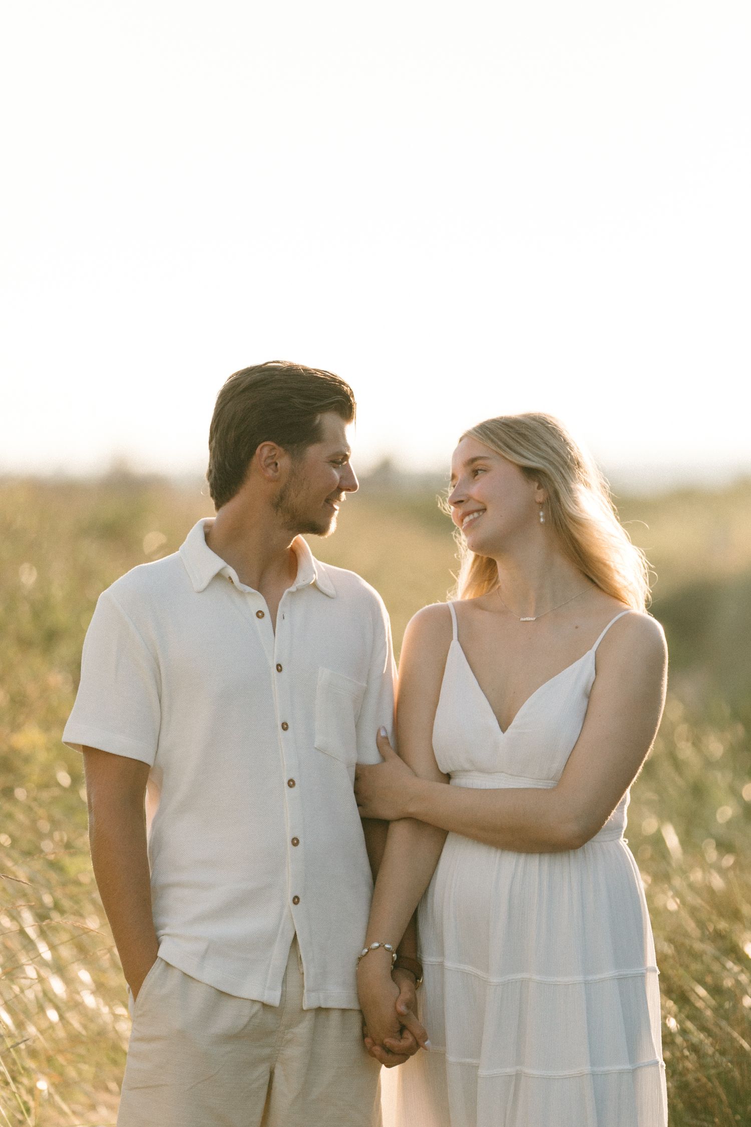 Romantic couple holding hands and smiling at each other in a sunlit golden field.