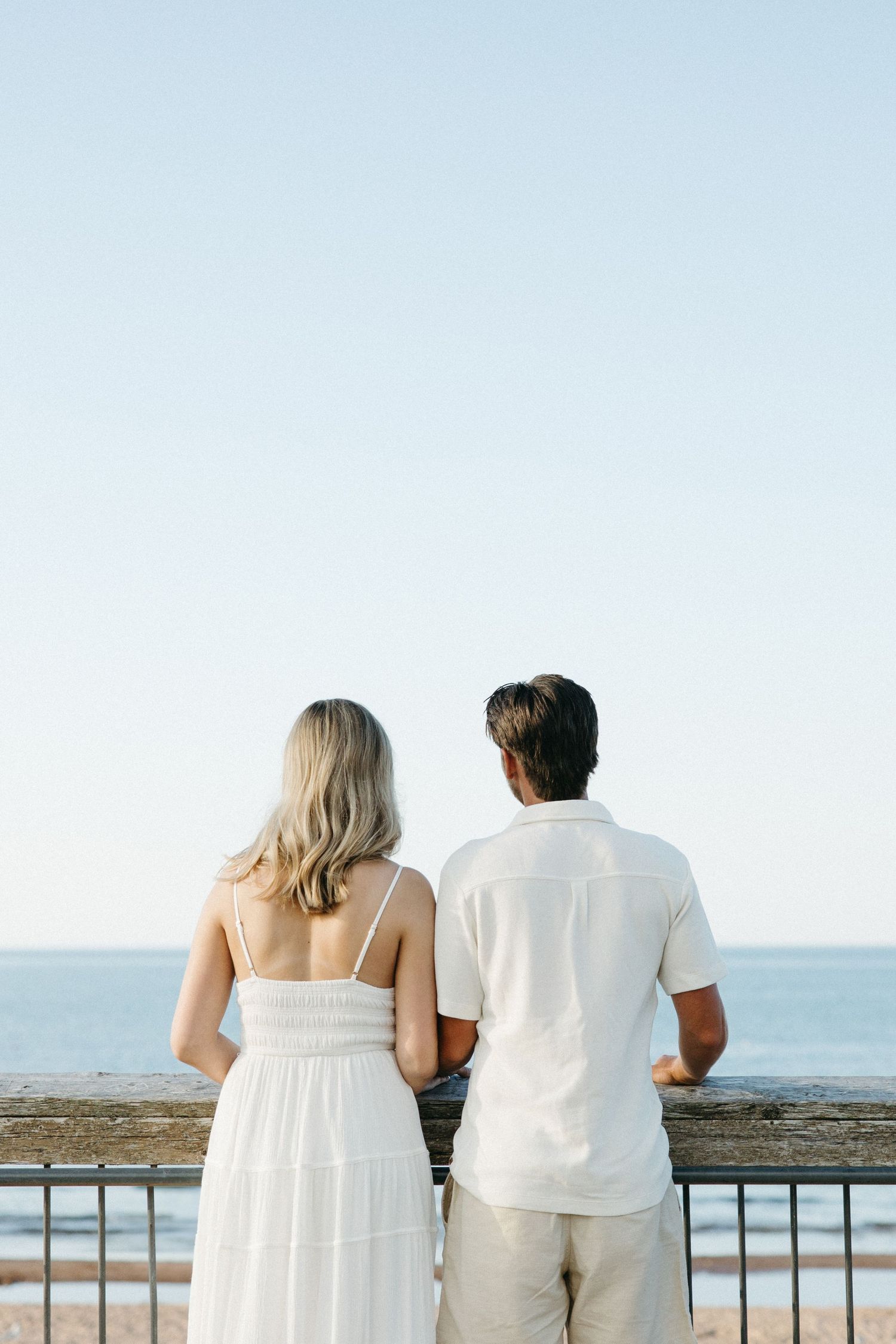 Couple in white outfits standing at a railing overlooking the ocean on a clear sunny day.