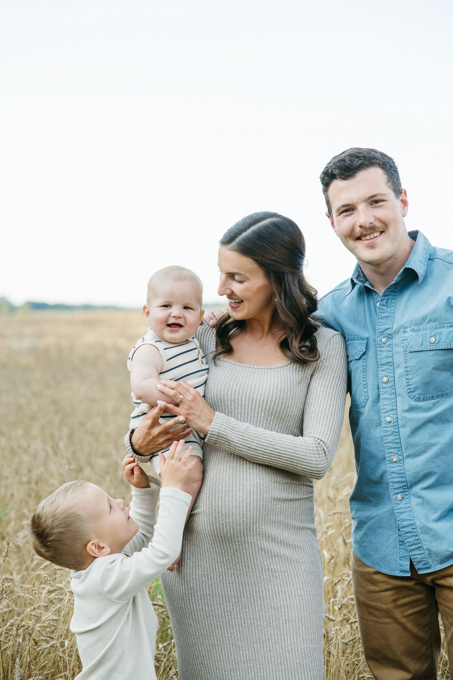 A happy pregnant woman holds a laughing baby while her husband and young son join her in a sunny wheat field.
