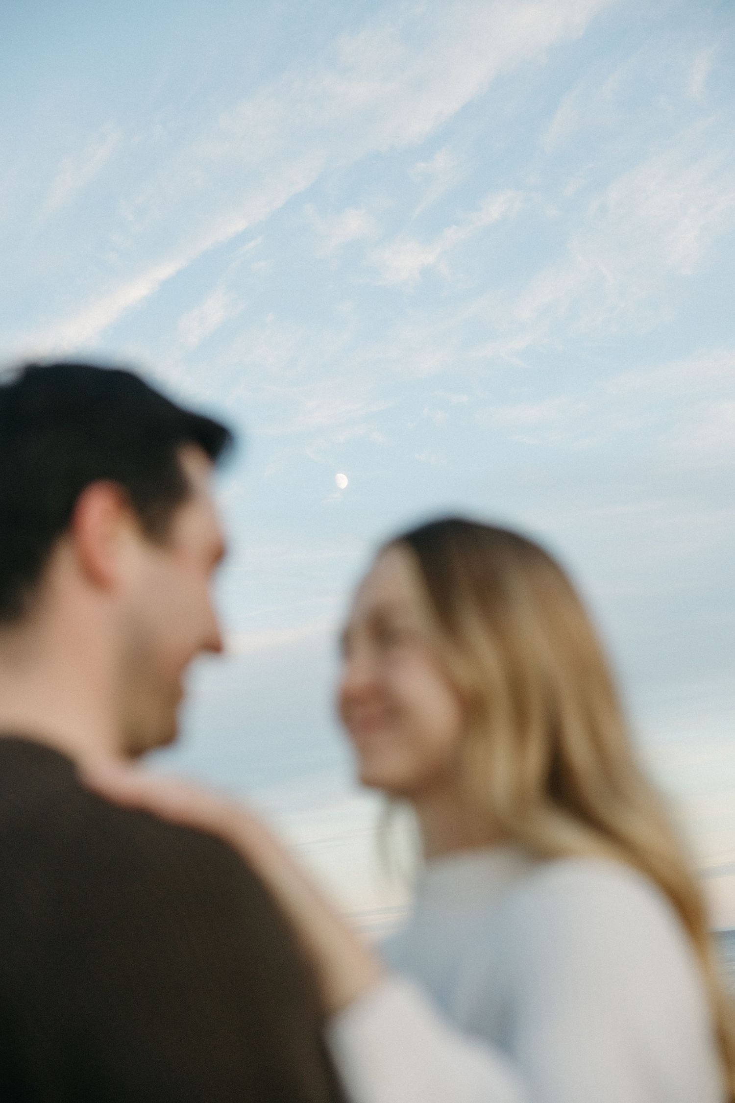 Blurred close-up of a smiling couple embracing outdoors under a soft blue sky with wispy clouds and a faint moon.
