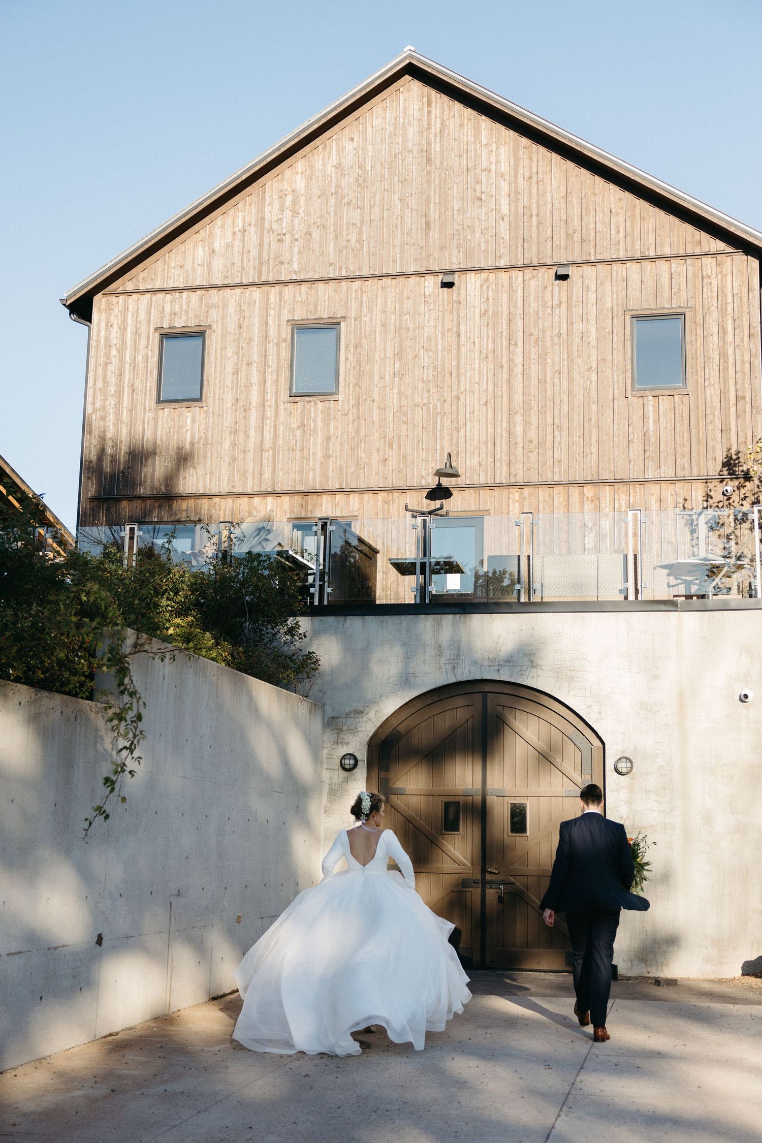 Newlyweds walk along a modern concrete wall with wooden barn backdrop creating artistic shadows.