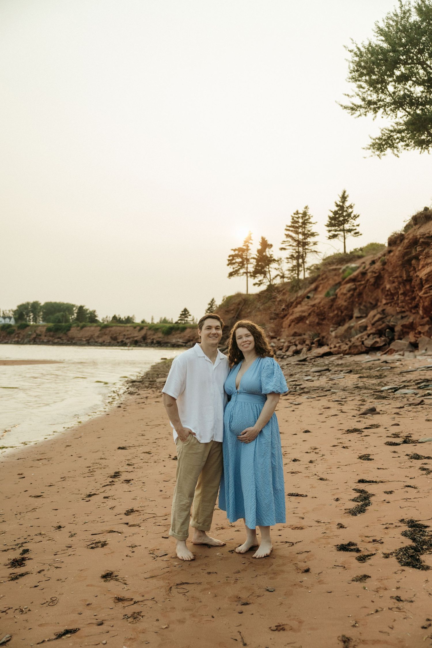 A couple poses together on a sandy beach with pine trees and cliffs in the background.