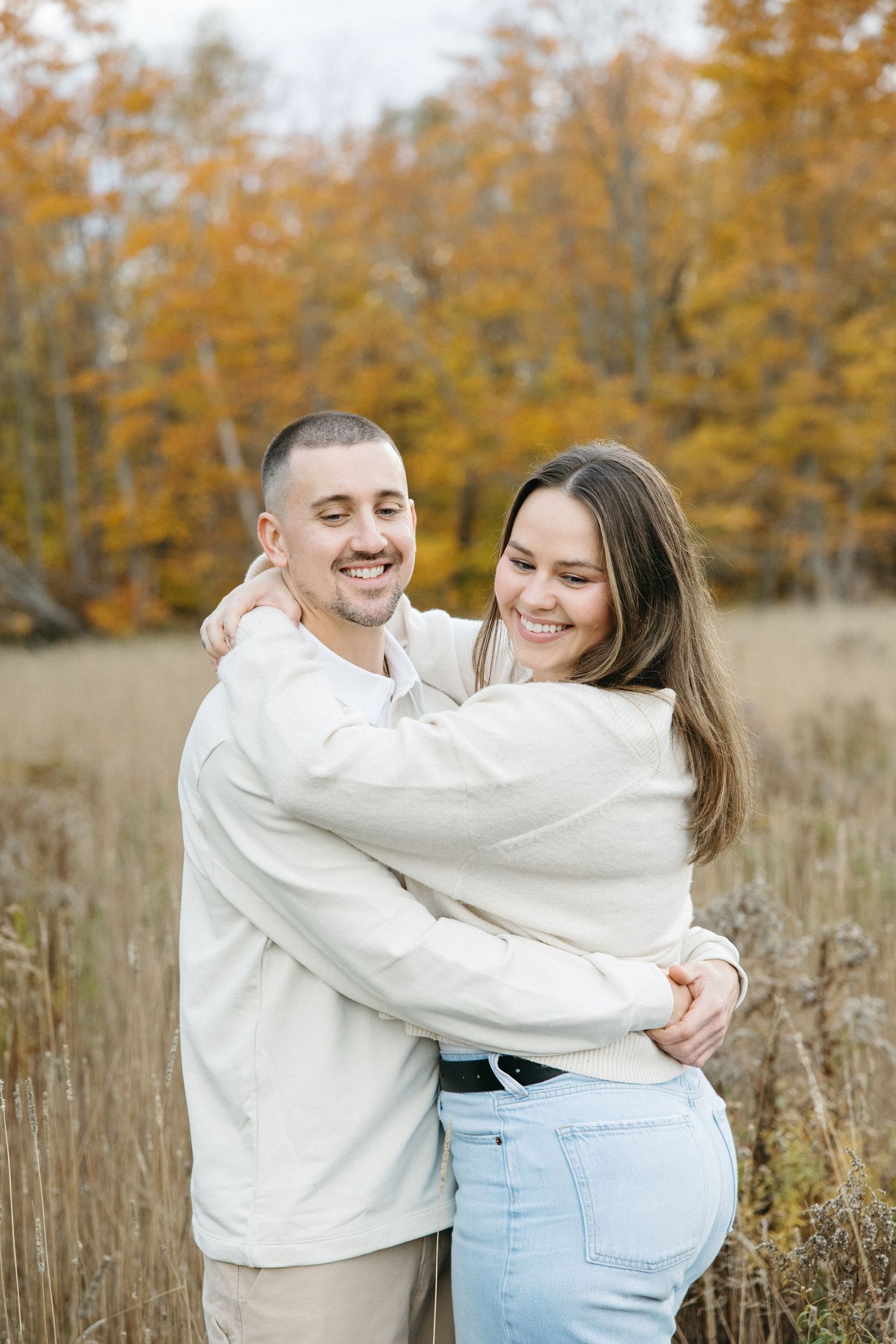 A happy couple in cream sweaters embrace and laugh together in a rustic autumn field.