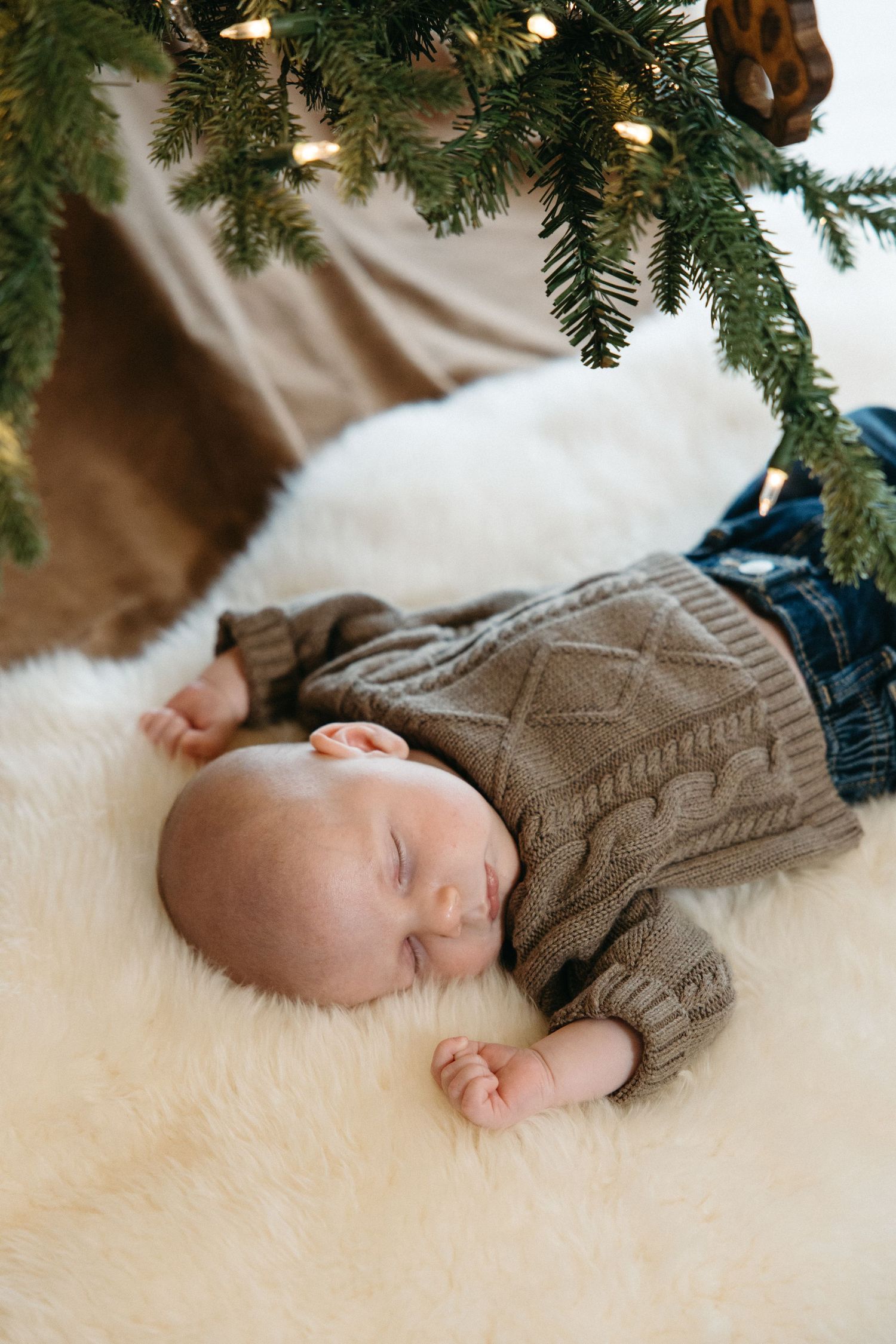 A sleeping infant wearing a brown cable knit sweater lies on a white fluffy rug beneath Christmas tree branches.