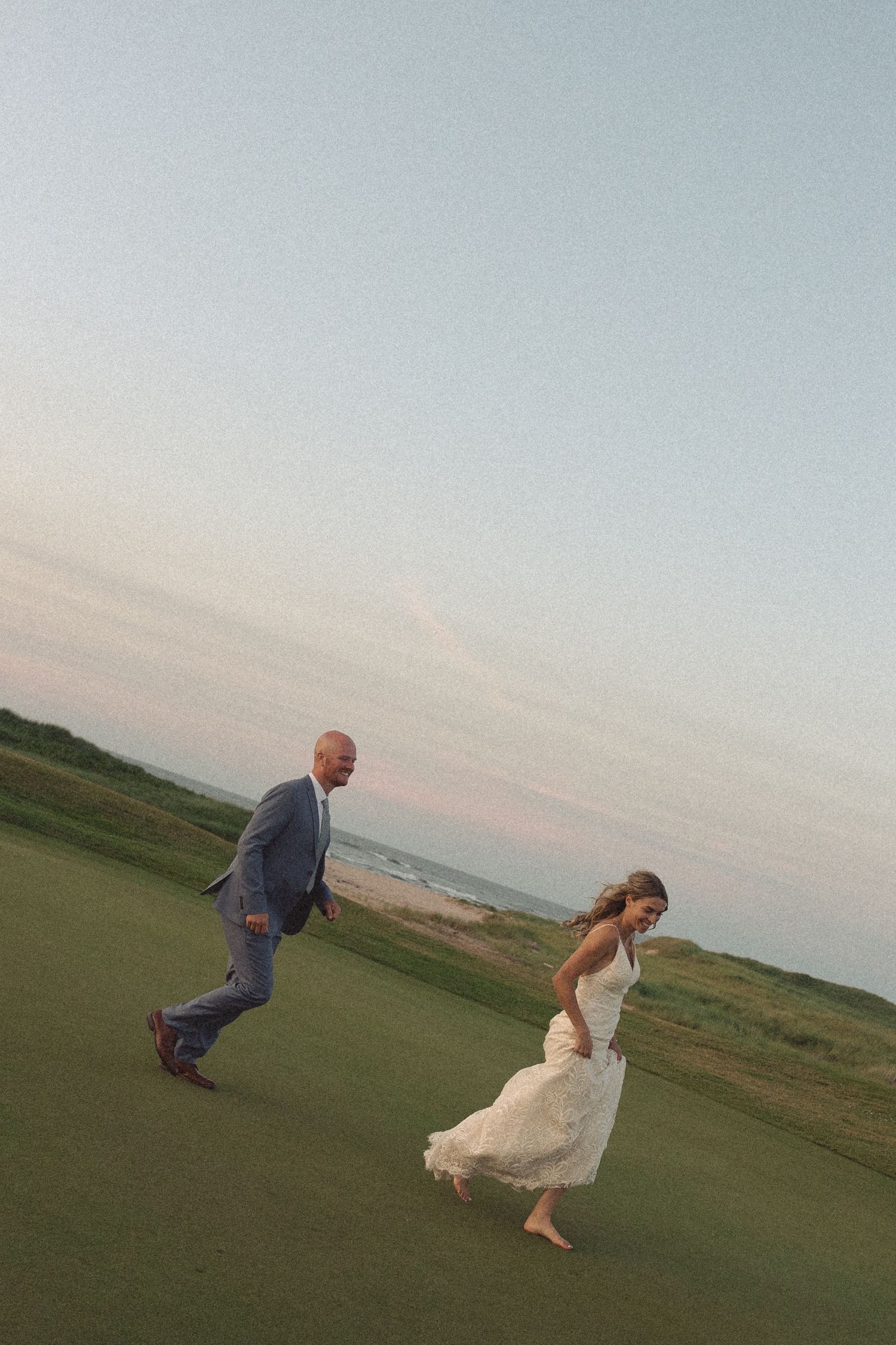 A couple playfully running and posing on a grassy hilltop overlooking the ocean.