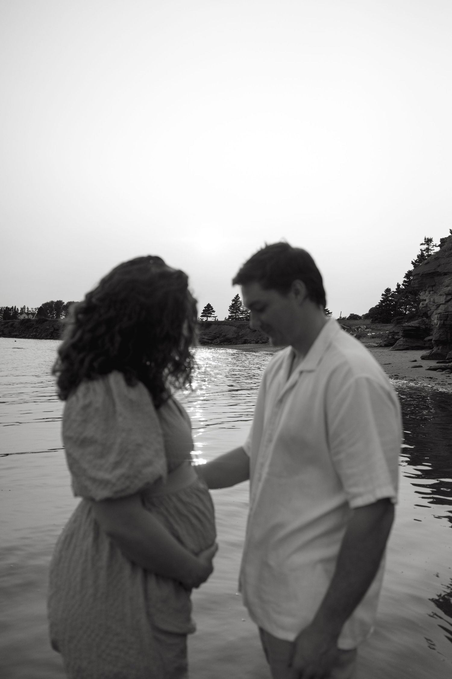 A couple shares playful moments on a beach at dusk with pine trees in the distance.