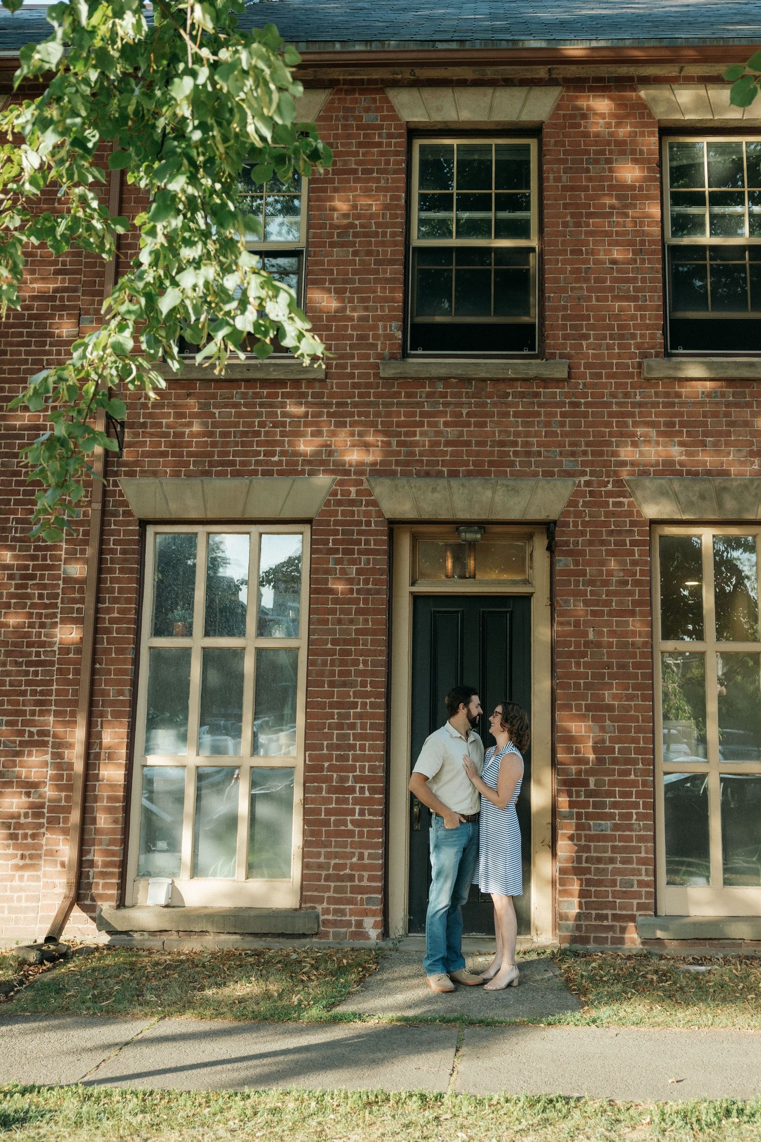A couple shares a tender moment in front of a brick building with large windows at sunset.