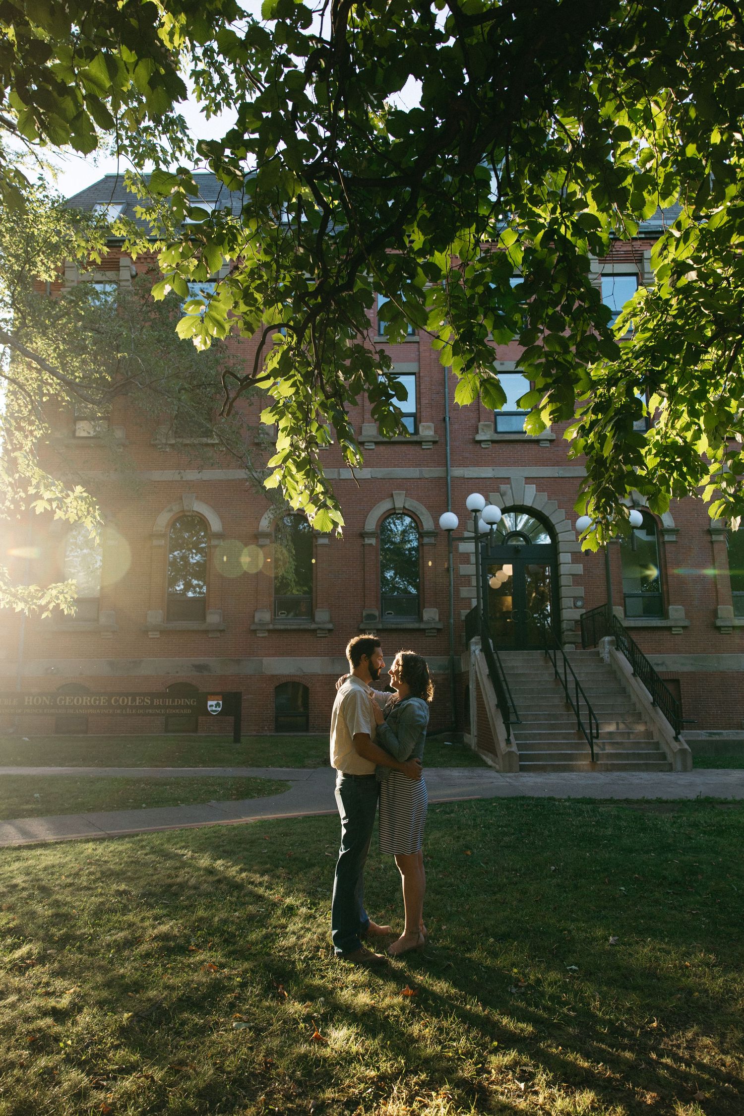 Two figures stand close together under green trees during sunset.