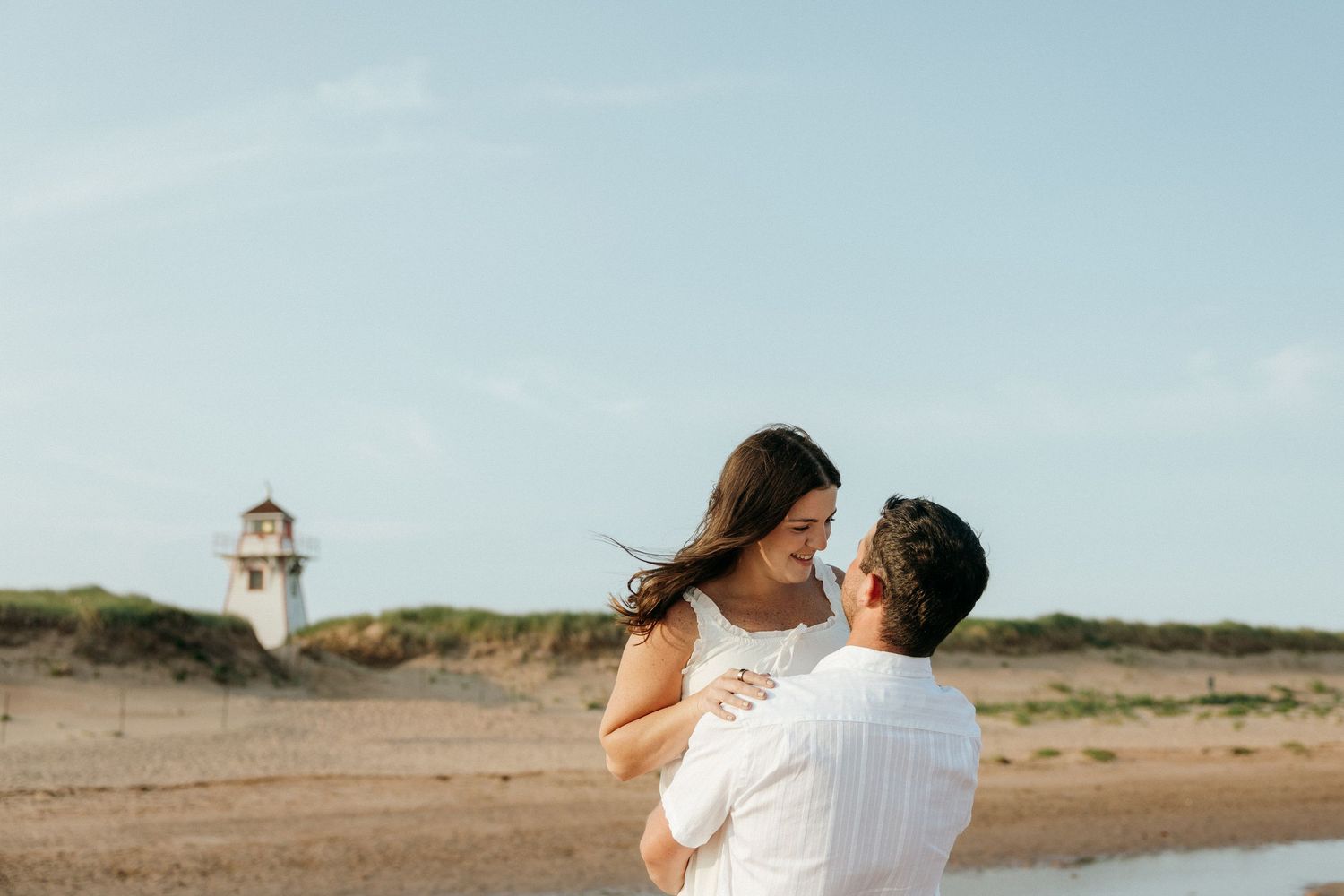 A romantic couple embraces on a sandy beach with a lighthouse visible in the background.