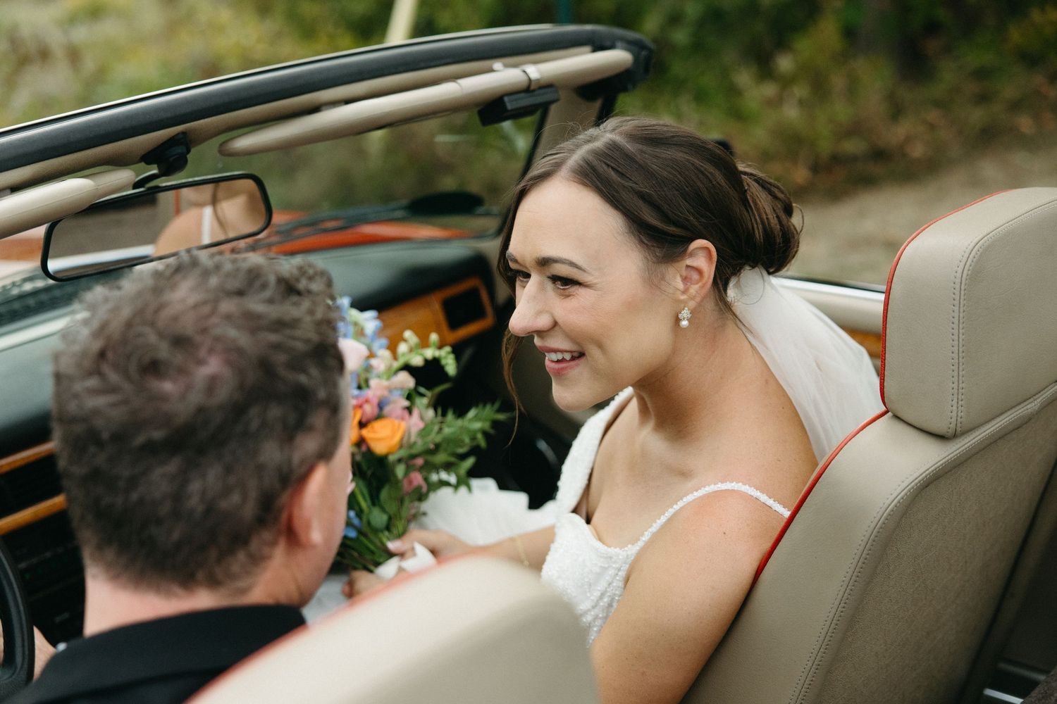 Couple sitting in vintage car with beige interior during wedding celebration.