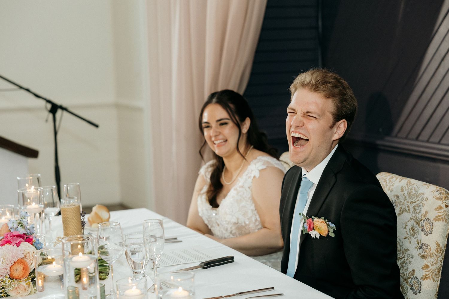 Newlyweds share joyful laughter while seated at their reception table.