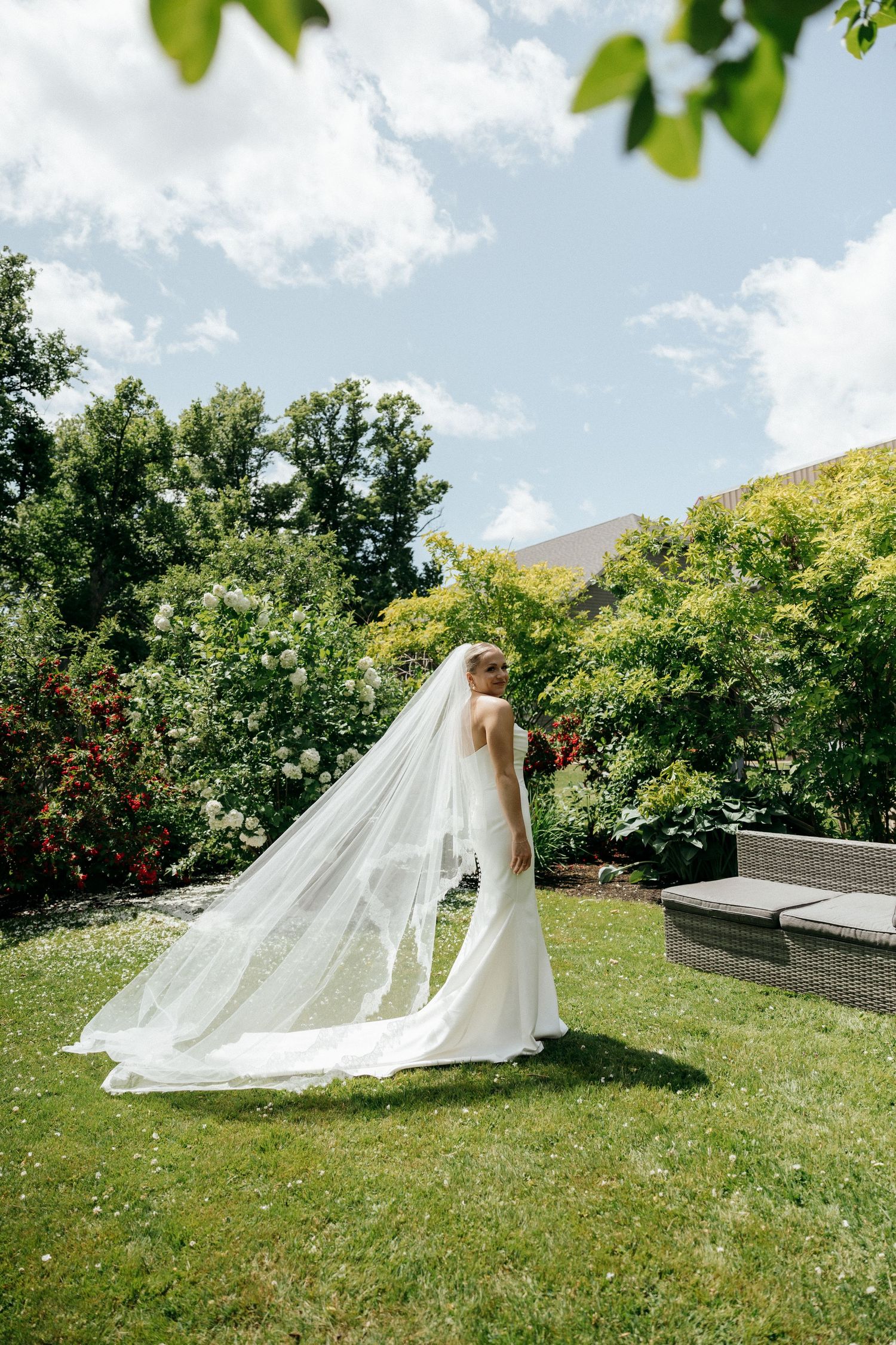 Bride in flowing white dress and veil walking through a sunny garden.