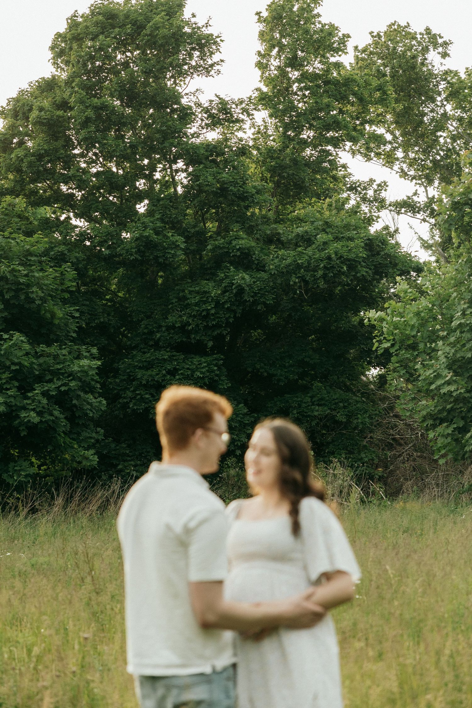 A couple in white clothing embraces while standing in a sunlit meadow with green trees in the background.