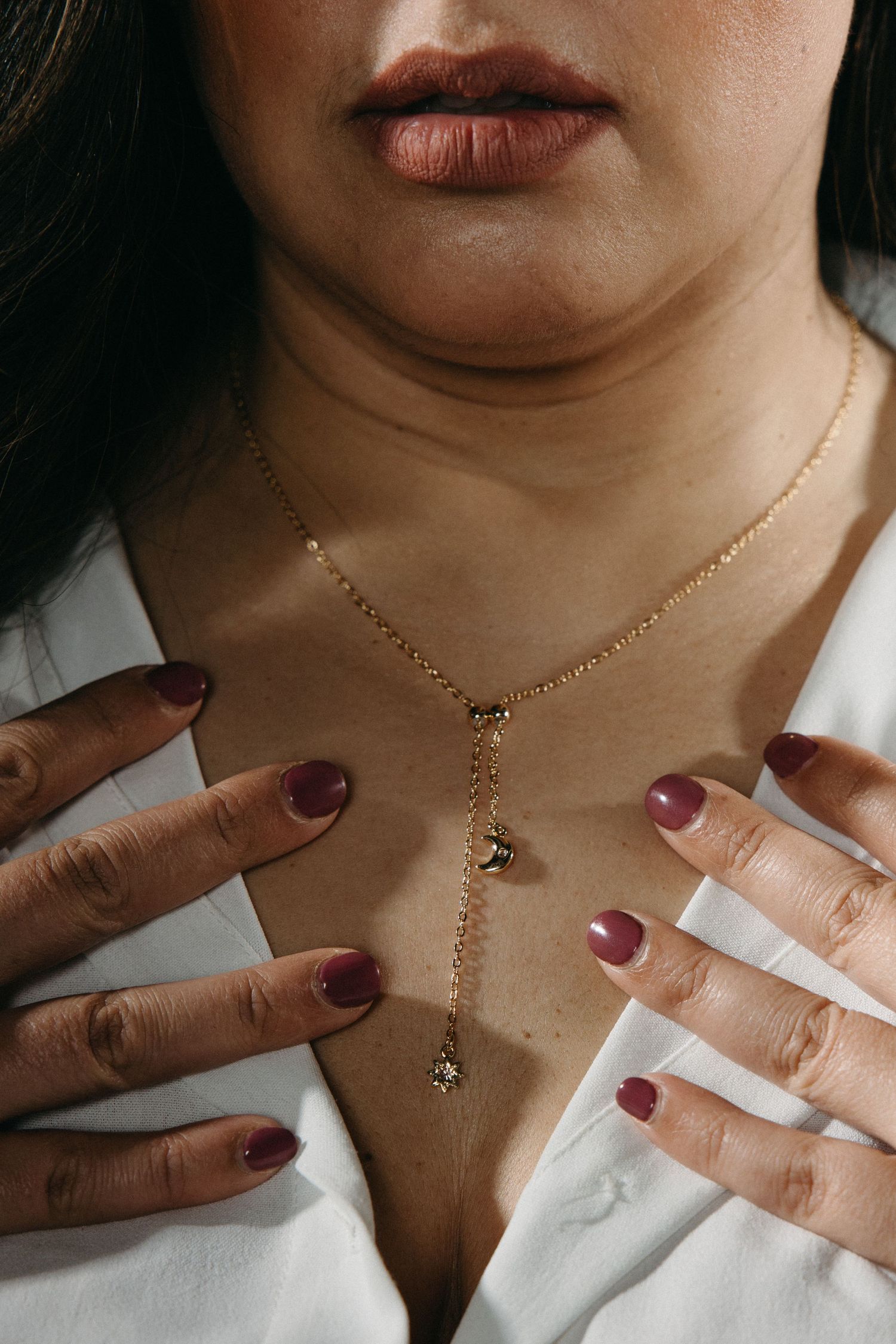 Close-up shots of hands with burgundy nails adjusting a delicate gold necklace against white clothing.