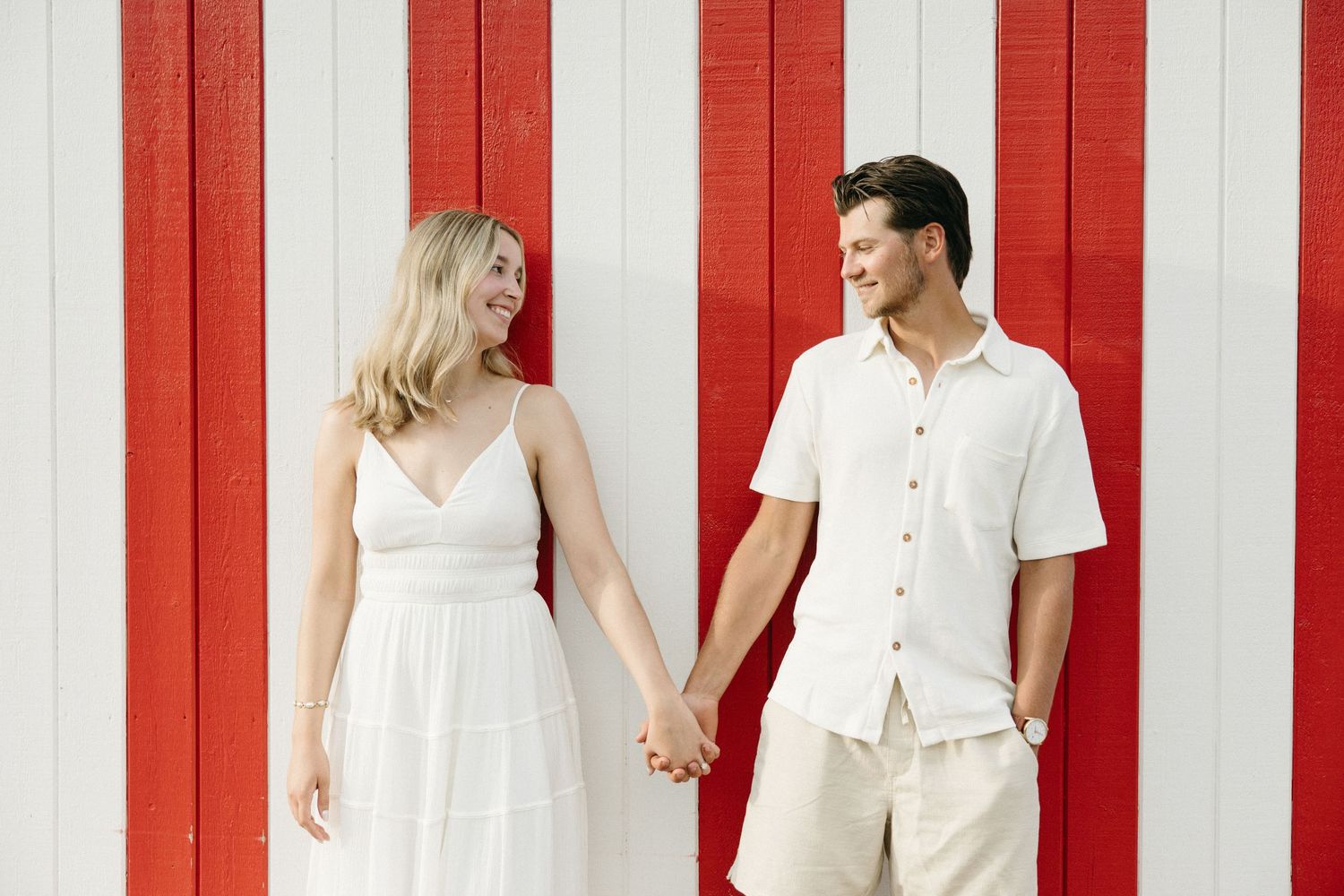A smiling couple holds hands in front of a red and white striped wall, woman in white dress, man in white shirt.