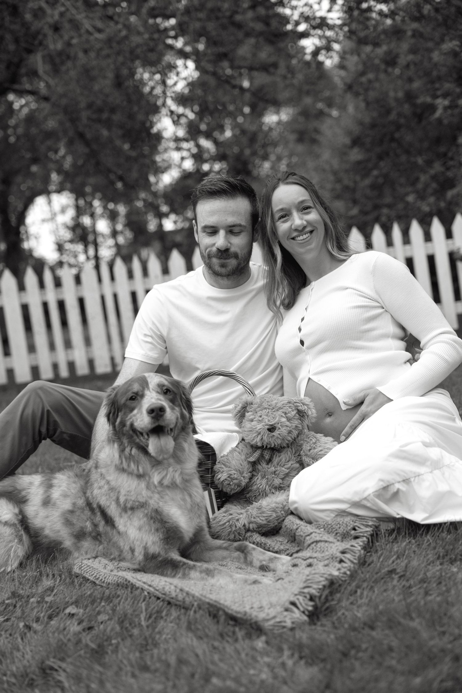 Black and white photo of smiling pregnant couple sitting on grass with their dog.