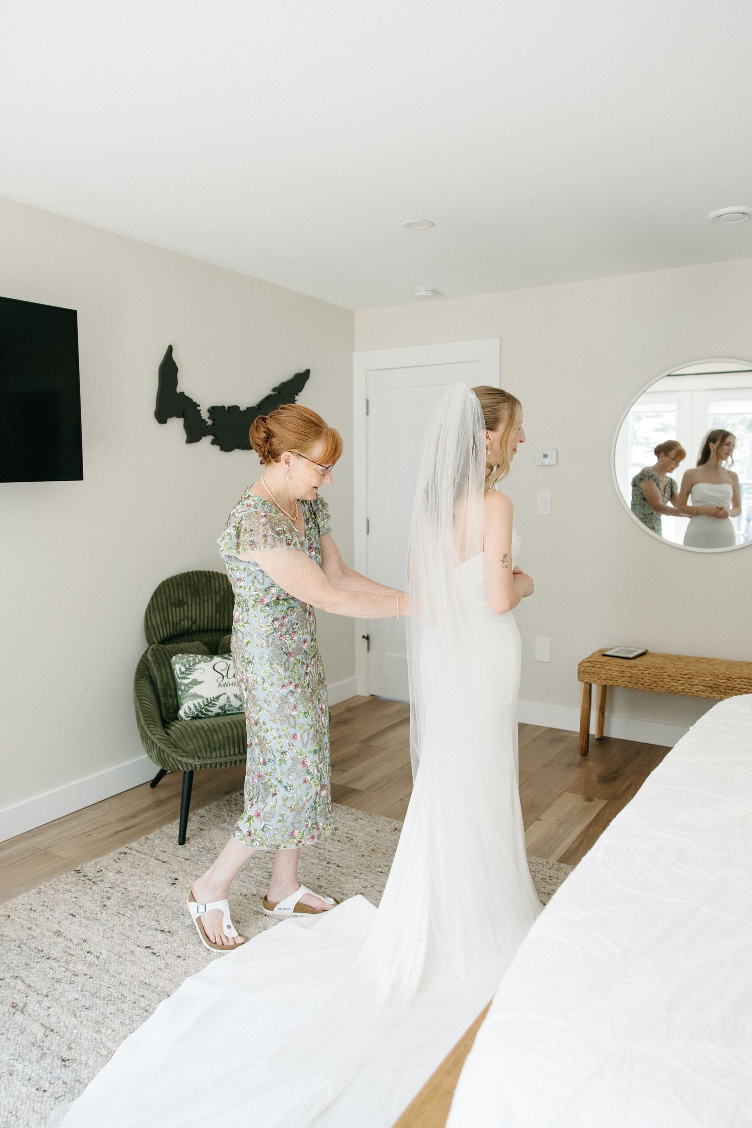 A bride in a white gown and veil gets help with her dress from a woman in a floral green dress in a bright bedroom.