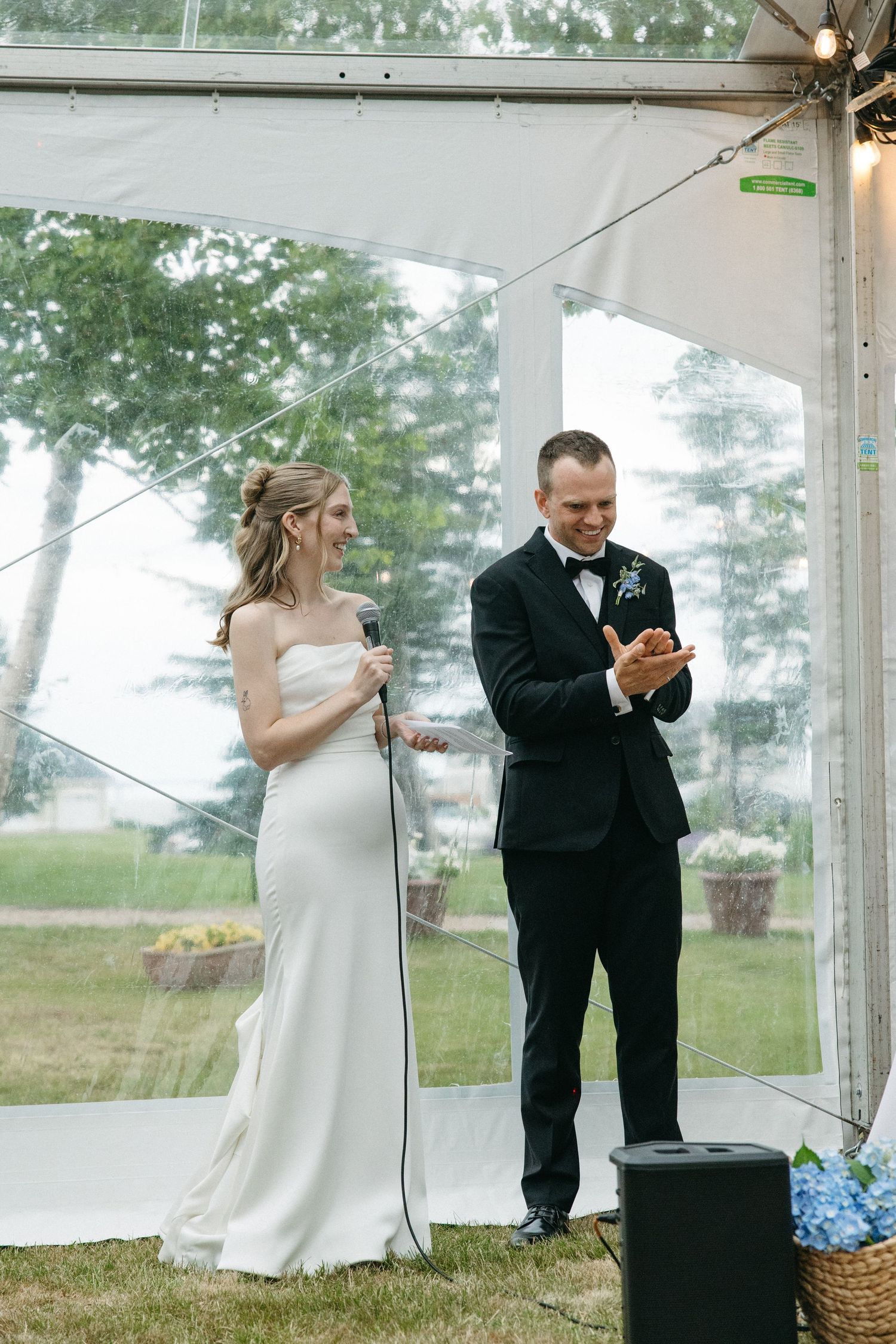 Bride holding microphone and groom in tuxedo speaking at wedding reception under clear tent.