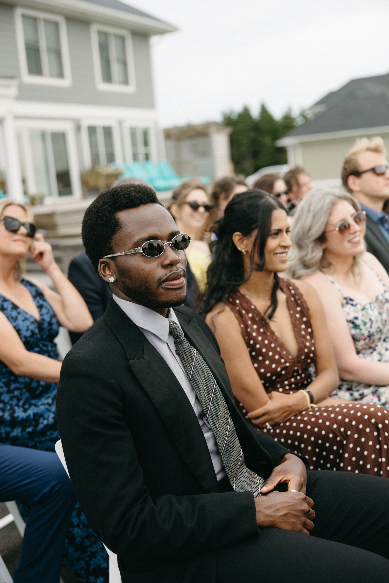 Stylish wedding guest in black suit and sunglasses seated at outdoor ceremony with diverse crowd behind him.