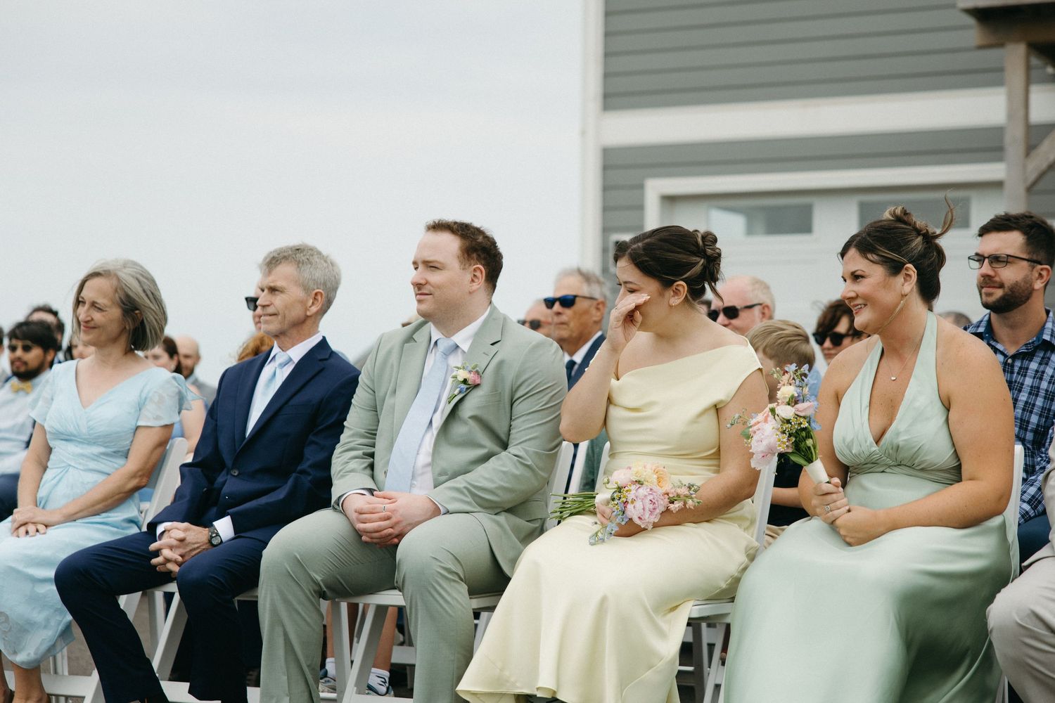 Wedding guests laughing and smiling while seated at outdoor ceremony including bridesmaids in pastel dresses.