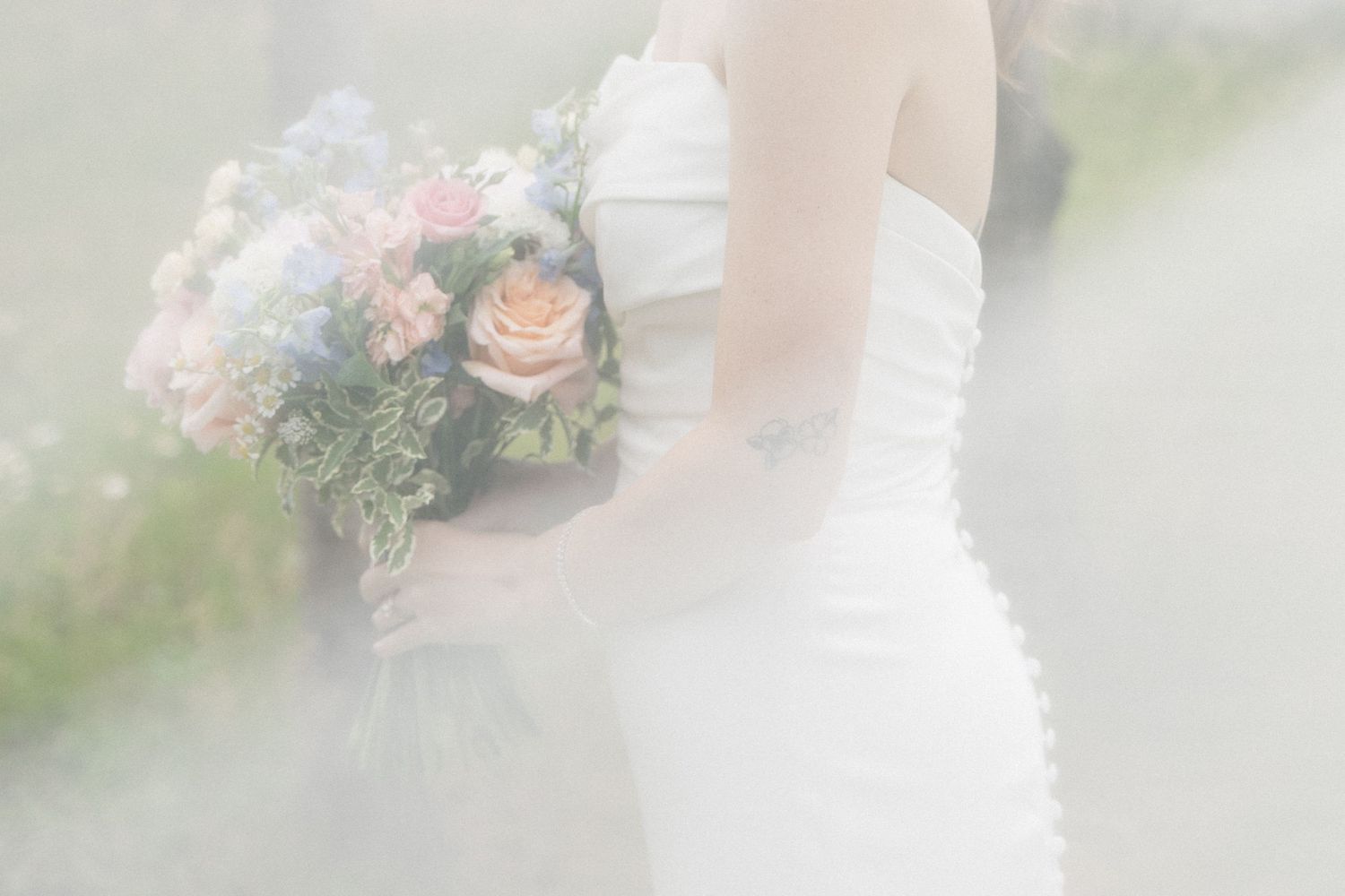 Bride in strapless white wedding gown holding lush pink and blue floral bouquet in dreamy soft-focus setting.