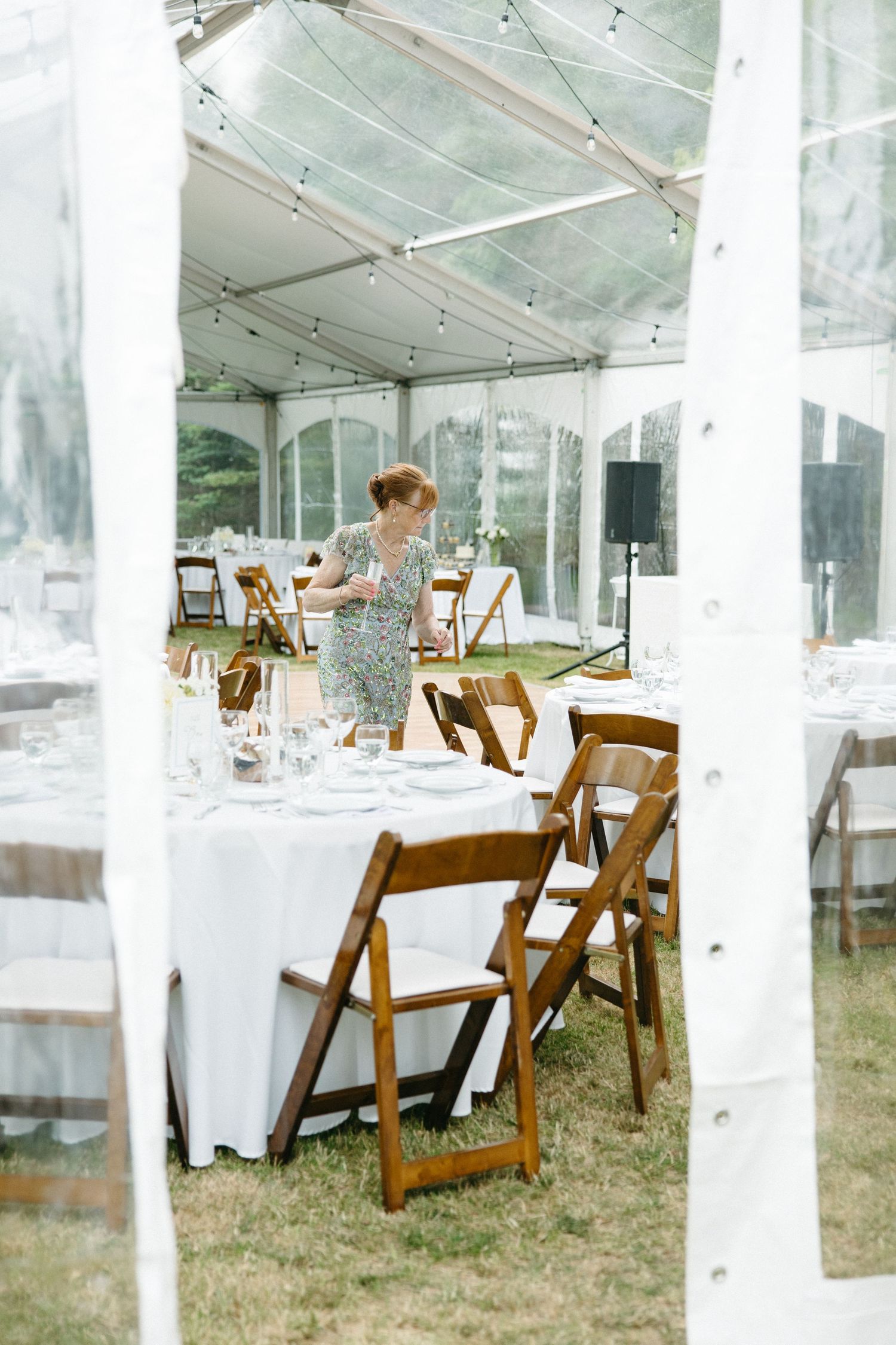 Elegant wedding reception setup inside a clear tent with white-draped round tables and wooden folding chairs.