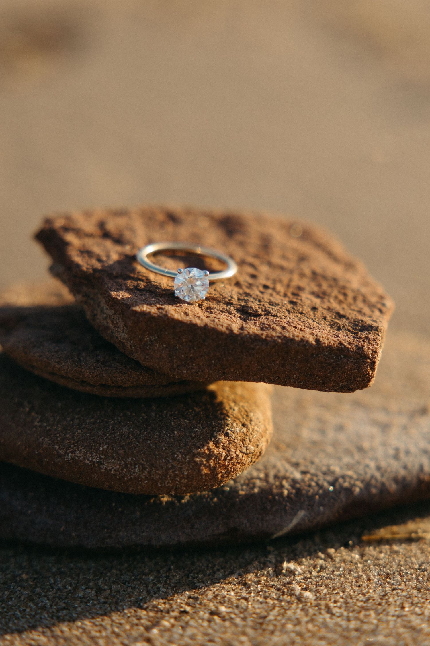 Diamond solitaire engagement ring resting on stacked rustic brown rocks on a sandy beach.