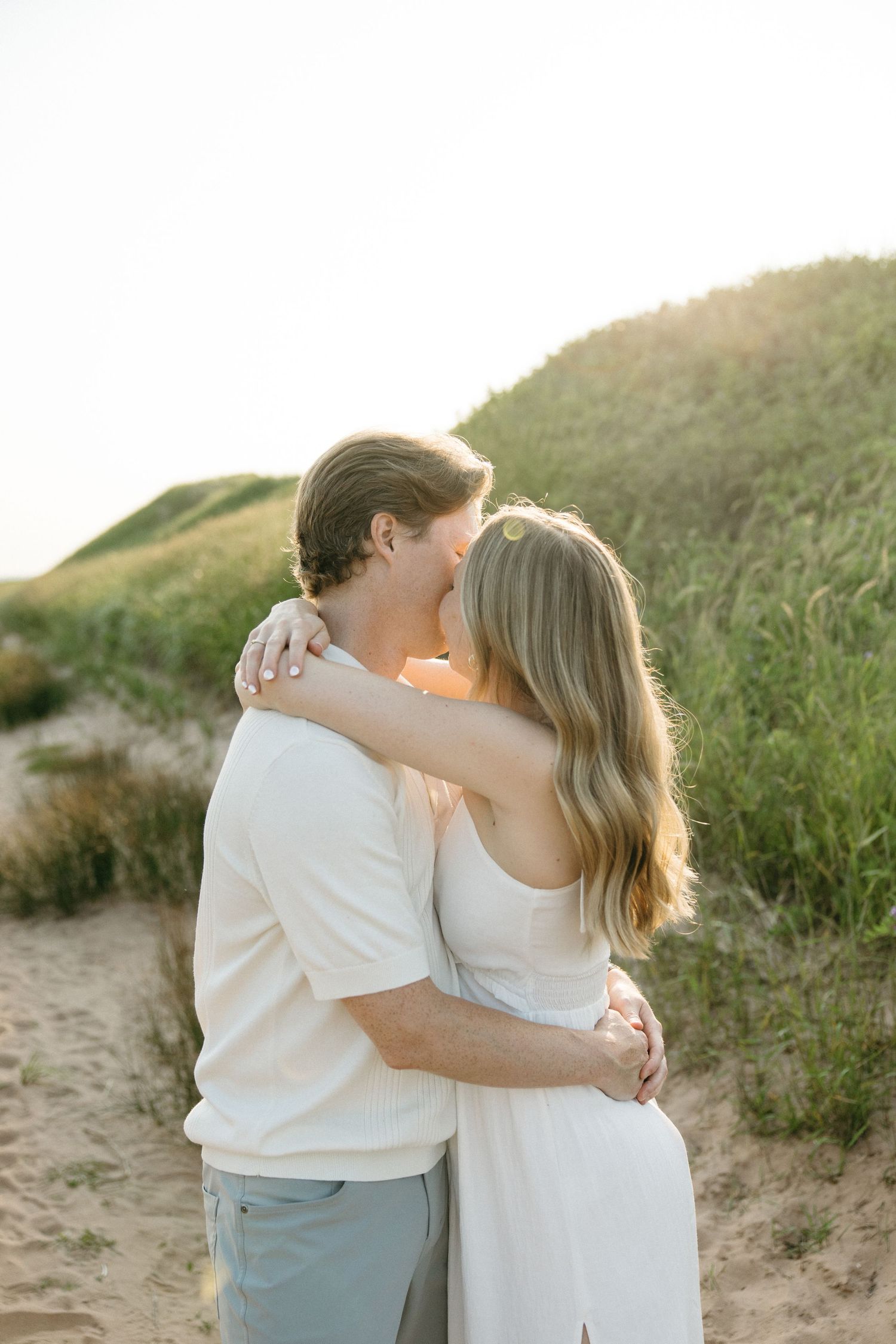 Couple sharing a romantic kiss among sandy dunes and green grass in warm golden hour light.