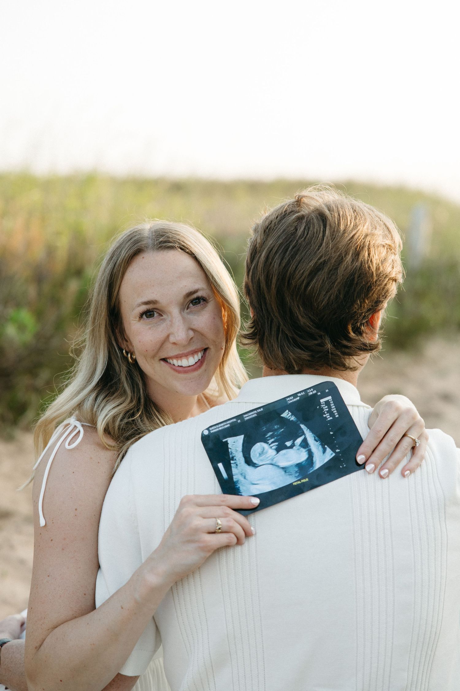 Smiling pregnant woman hugging man on beach path while holding ultrasound photo behind his back.