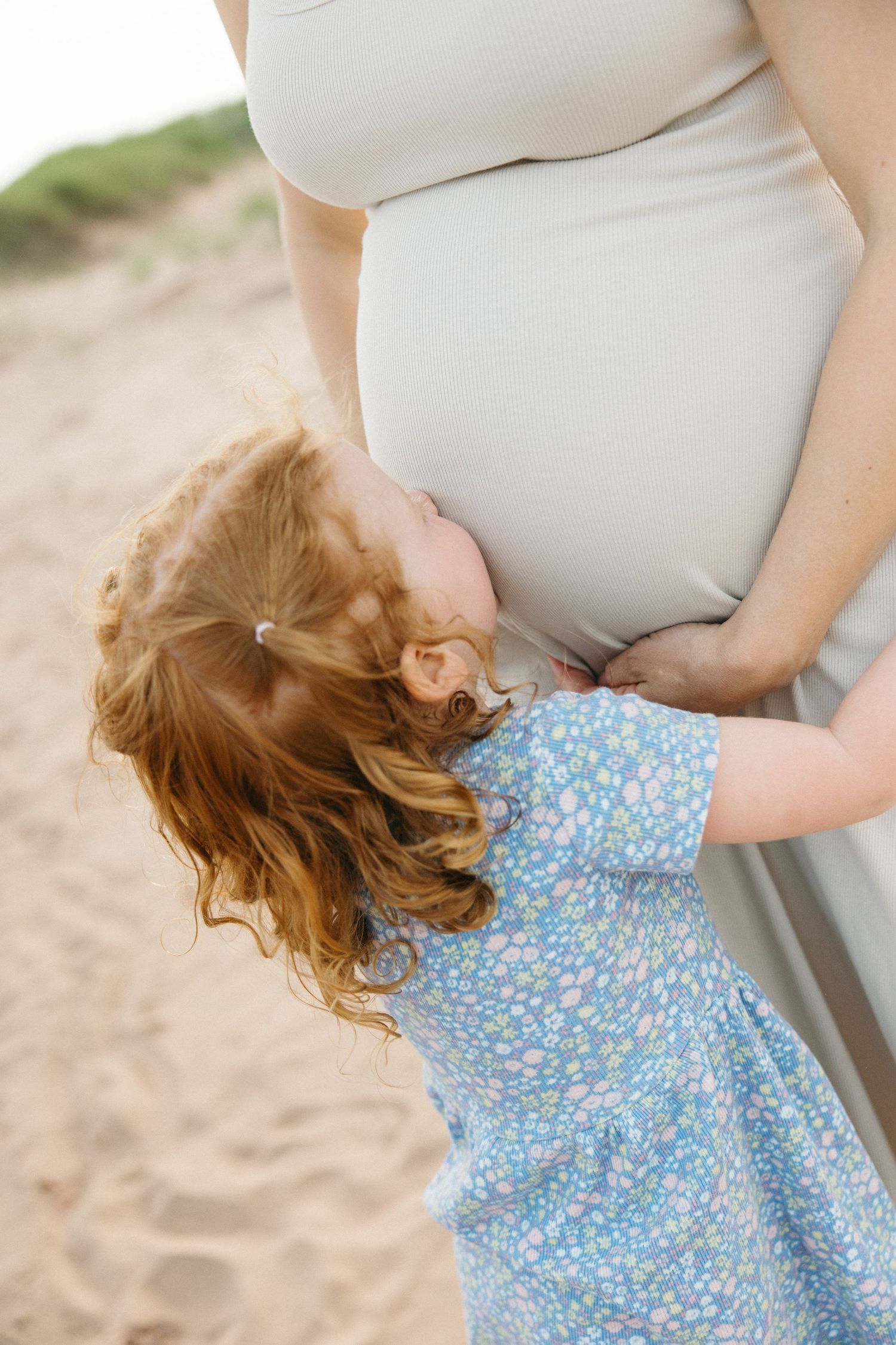 A red-haired toddler girl in a floral dress hugs a pregnant womans belly on a sandy beach.