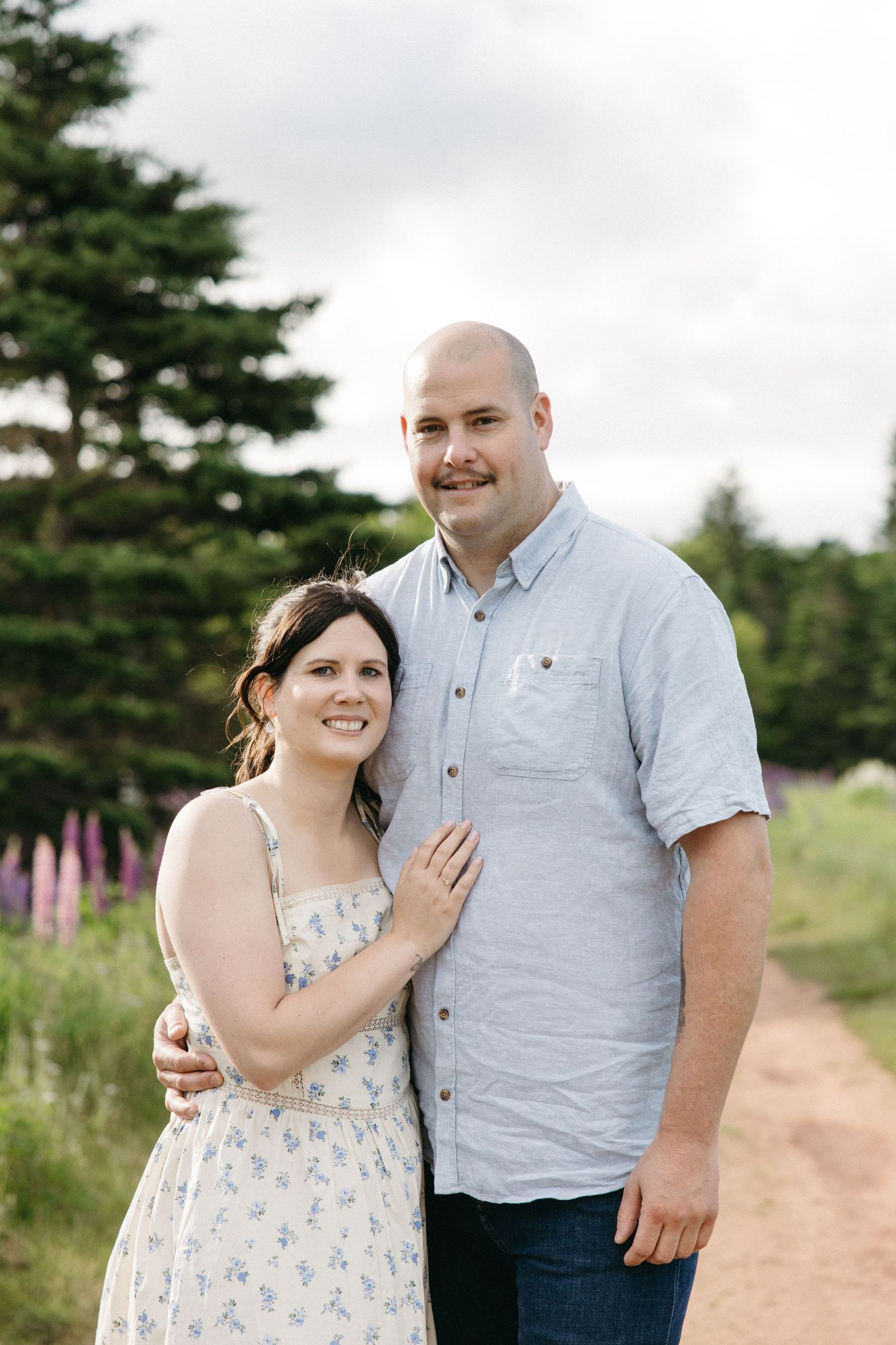 Smiling couple posing outdoors on a dirt path surrounded by green trees and wildflowers on a cloudy day.