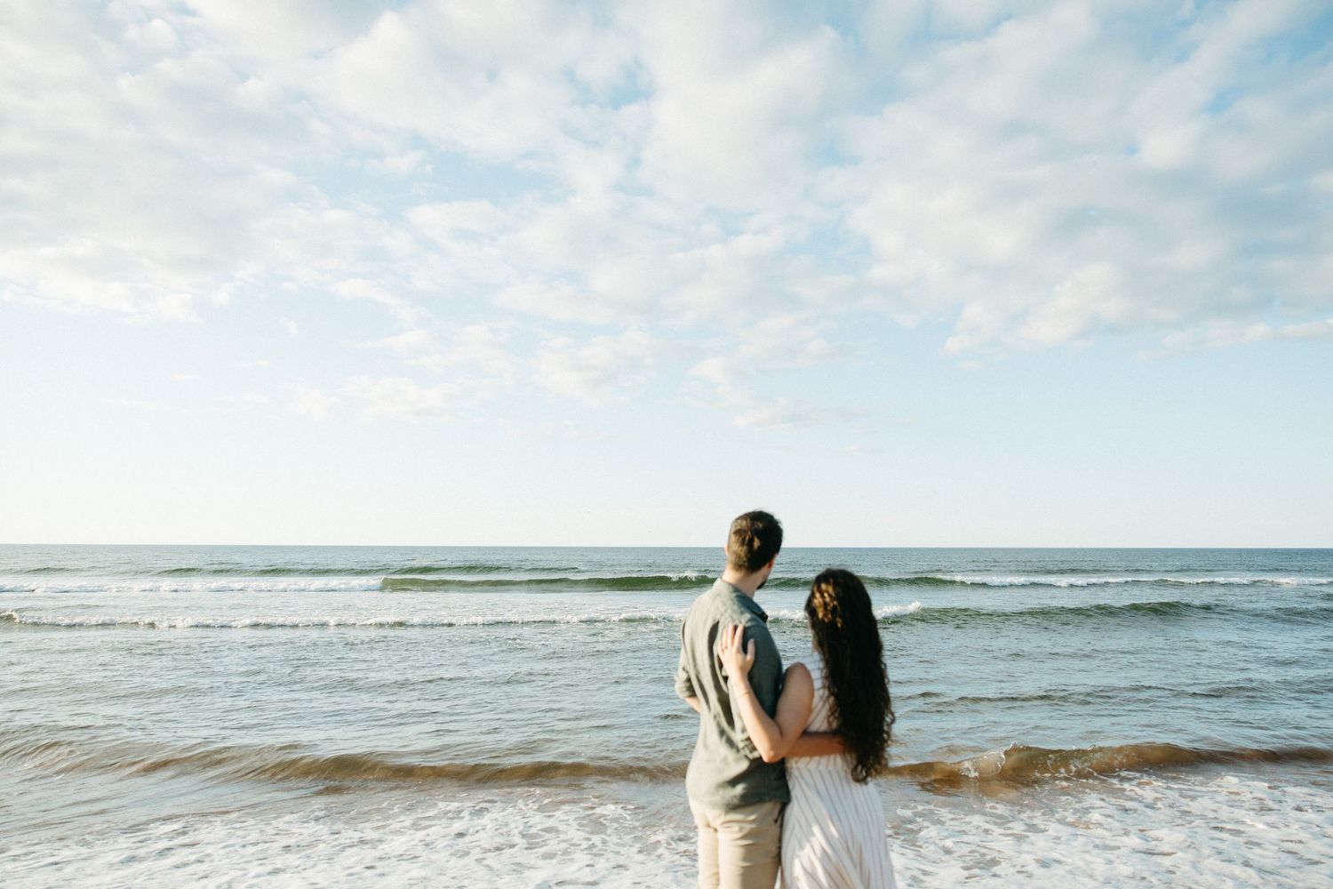 Couple standing together on a sandy beach gazing at ocean waves under a partly cloudy sky.
