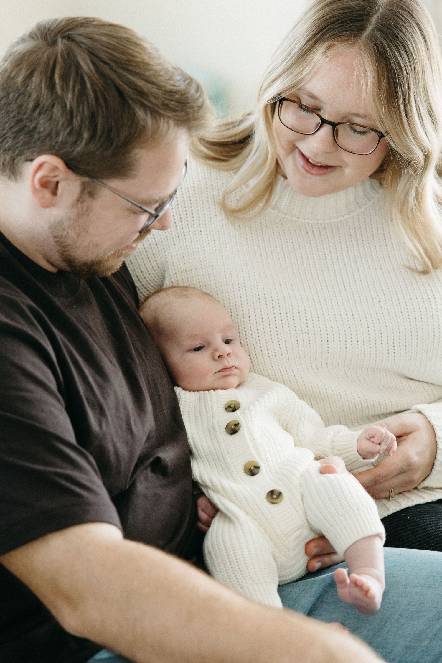 A couple in cozy sweaters cuddles with their newborn baby wearing a white knit outfit with wooden buttons.